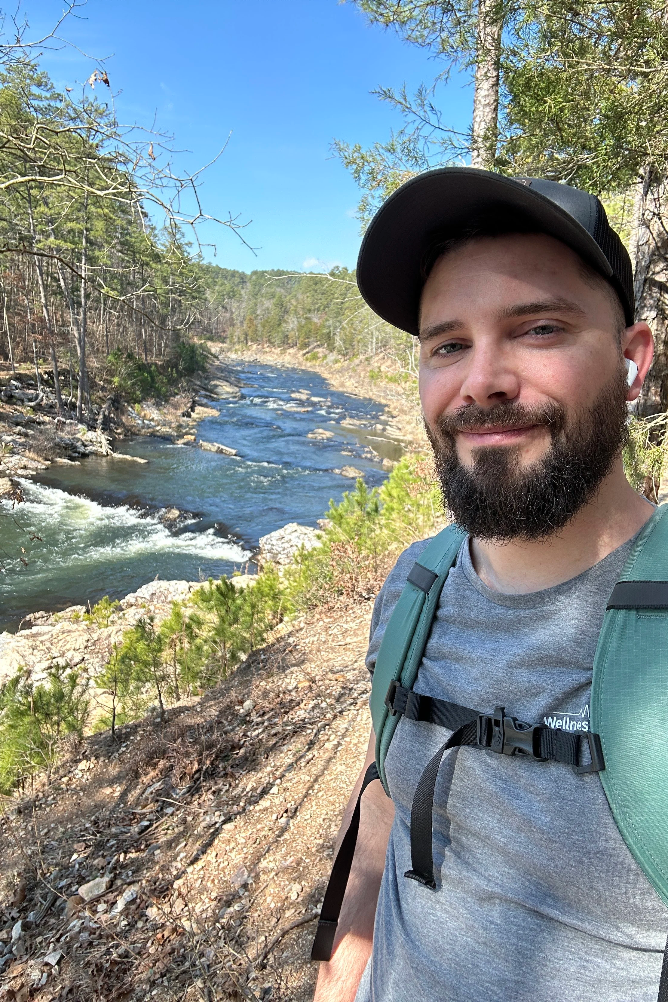 A man hiking along a river trail surrounded by trees under a blue sky.