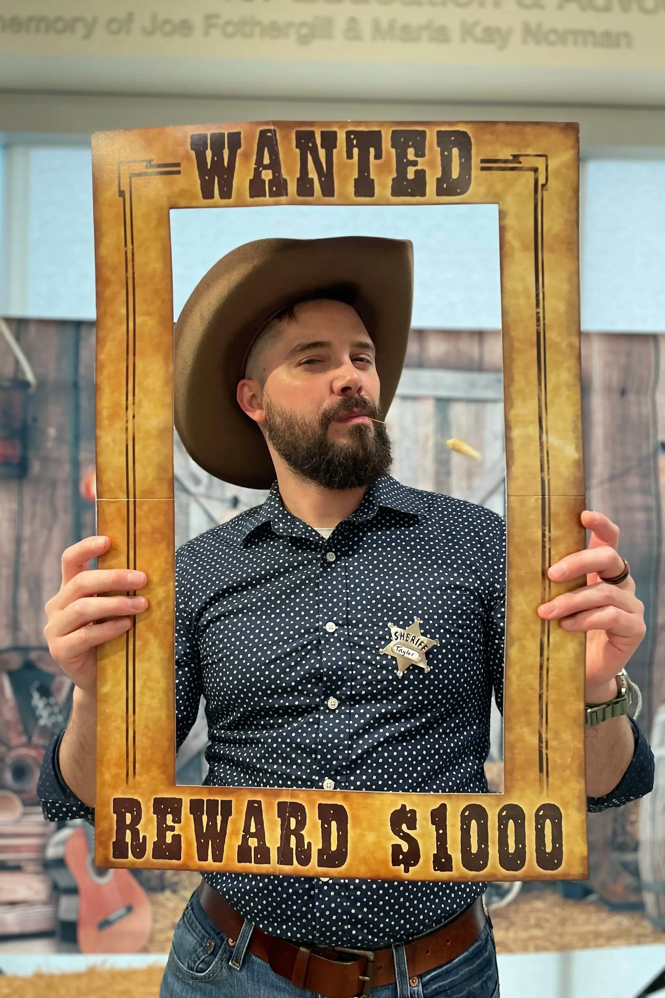 Man dressed as a cowboy sheriff holding a large, empty wanted poster frame with the words 'WANTED REWARD $1000' at the bottom. He's wearing a wide-brimmed cowboy hat, a polka-dot button-up shirt, and has a badge on his chest. The background shows a rustic wooden fence and guitars.