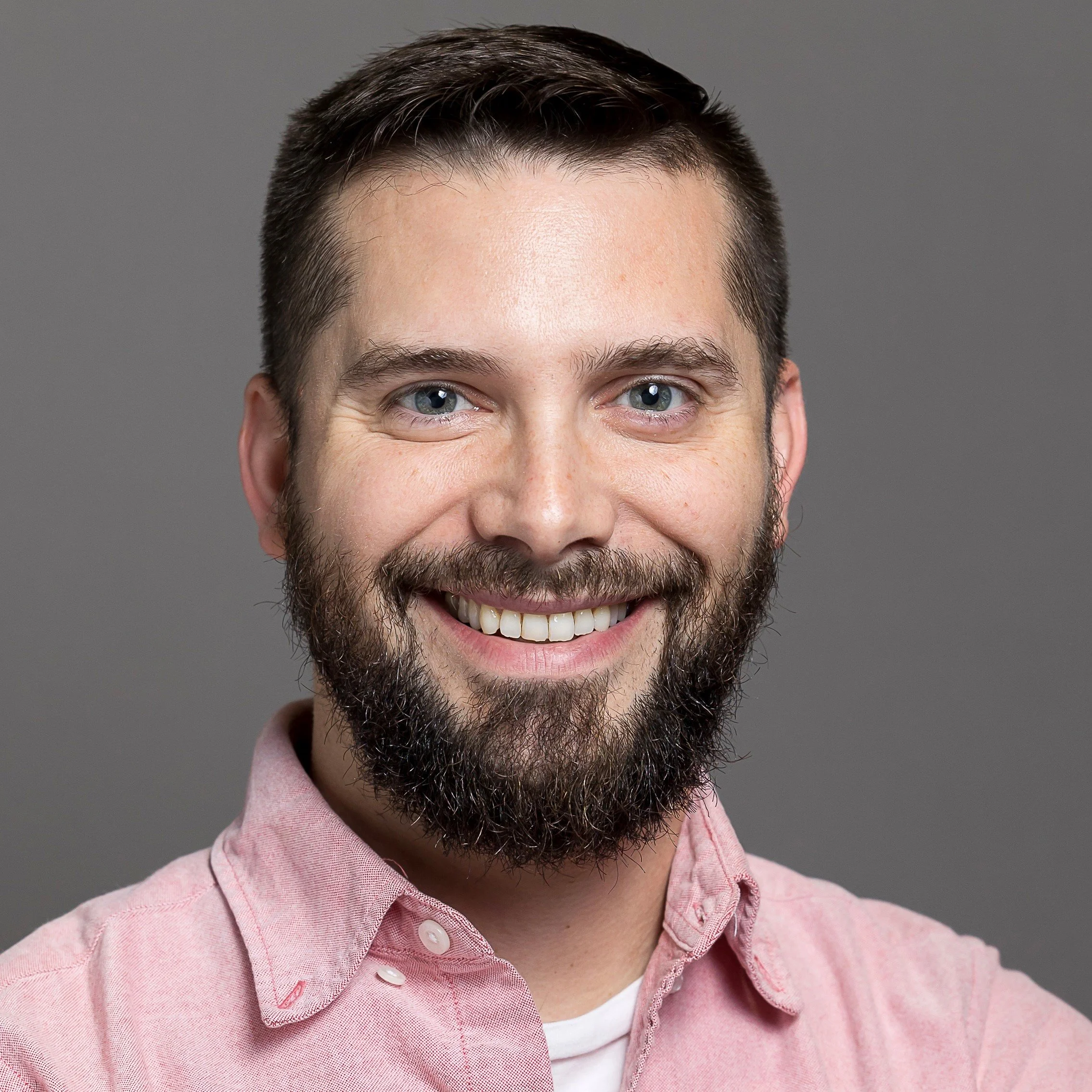 A smiling man with a beard and short dark hair wearing a pink collared shirt against a gray background.