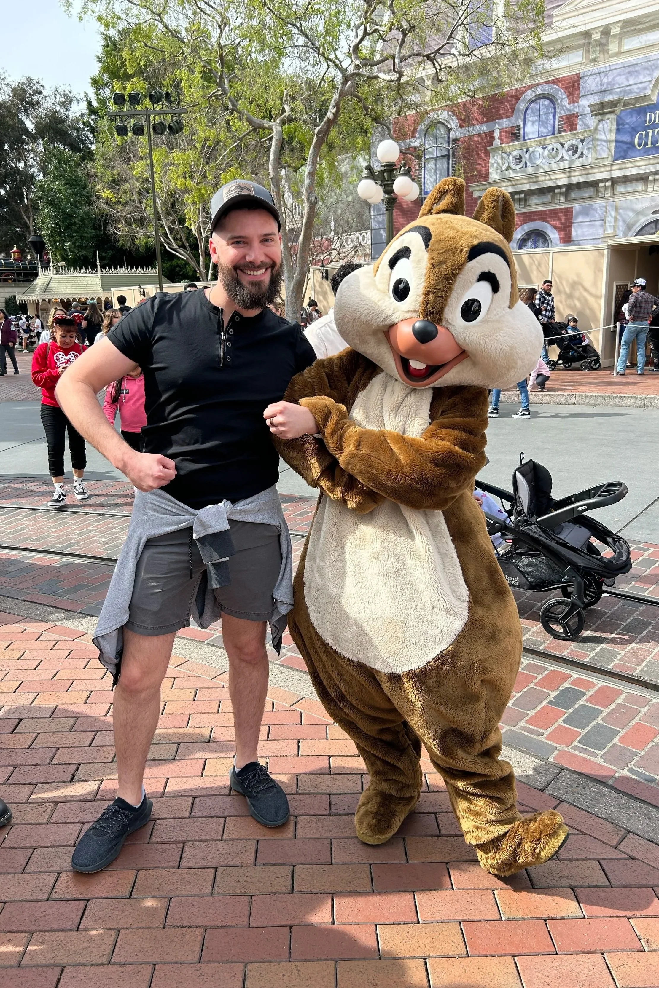 A man standing next to a person in a chipmunk costume at a theme park, smiling for the camera. The background shows other visitors, trees, and a building with decorative architecture.