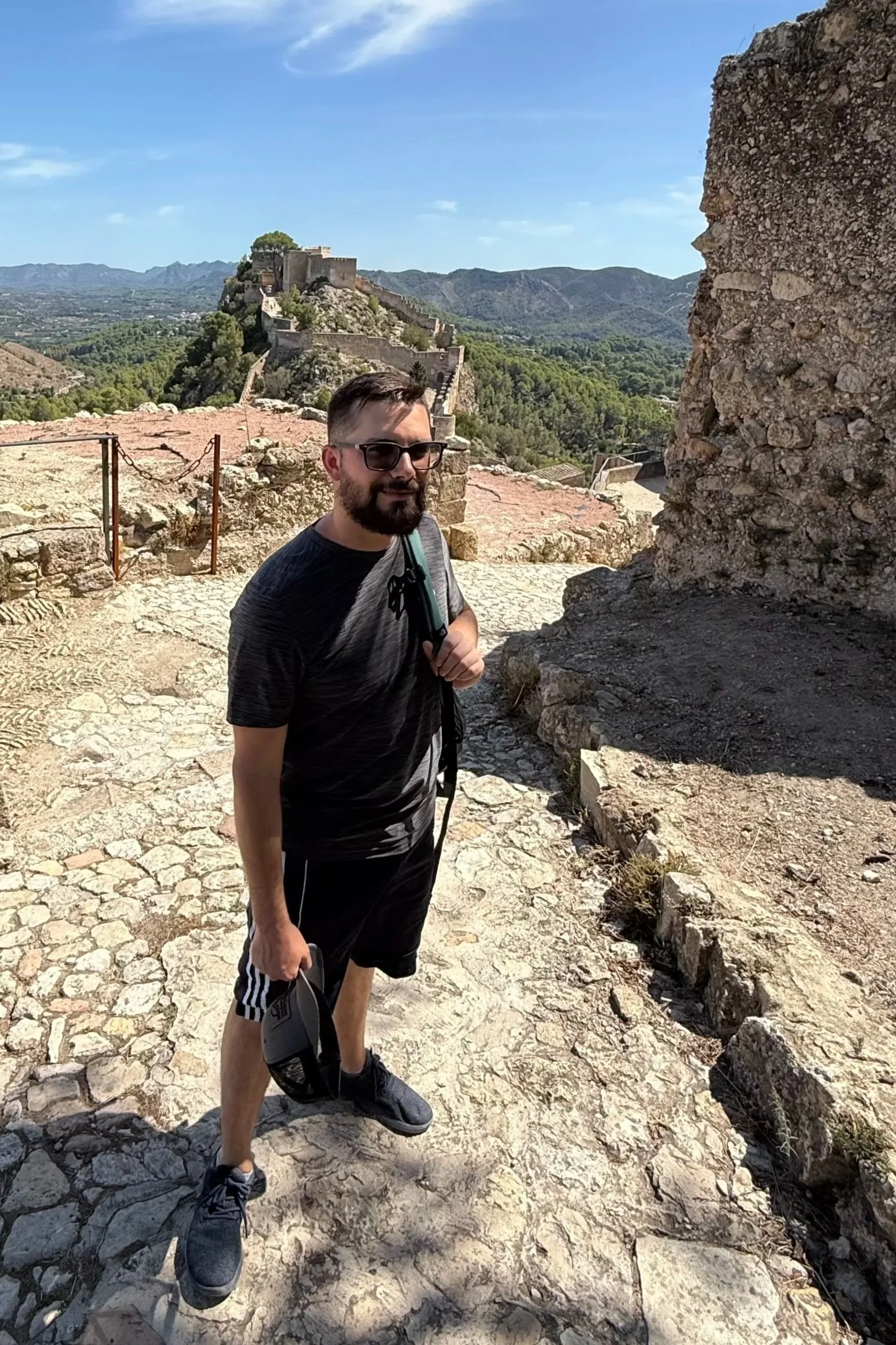 A man with sunglasses, a black and gray t-shirt, black shorts, and black shoes stands on a cobblestone path at a historical site with ancient stone ruins. He holds a hat in his right hand and has a backpack strap over his left shoulder. In the background, there is a fortress on a hill, greenery, and a mountain range under a blue sky.