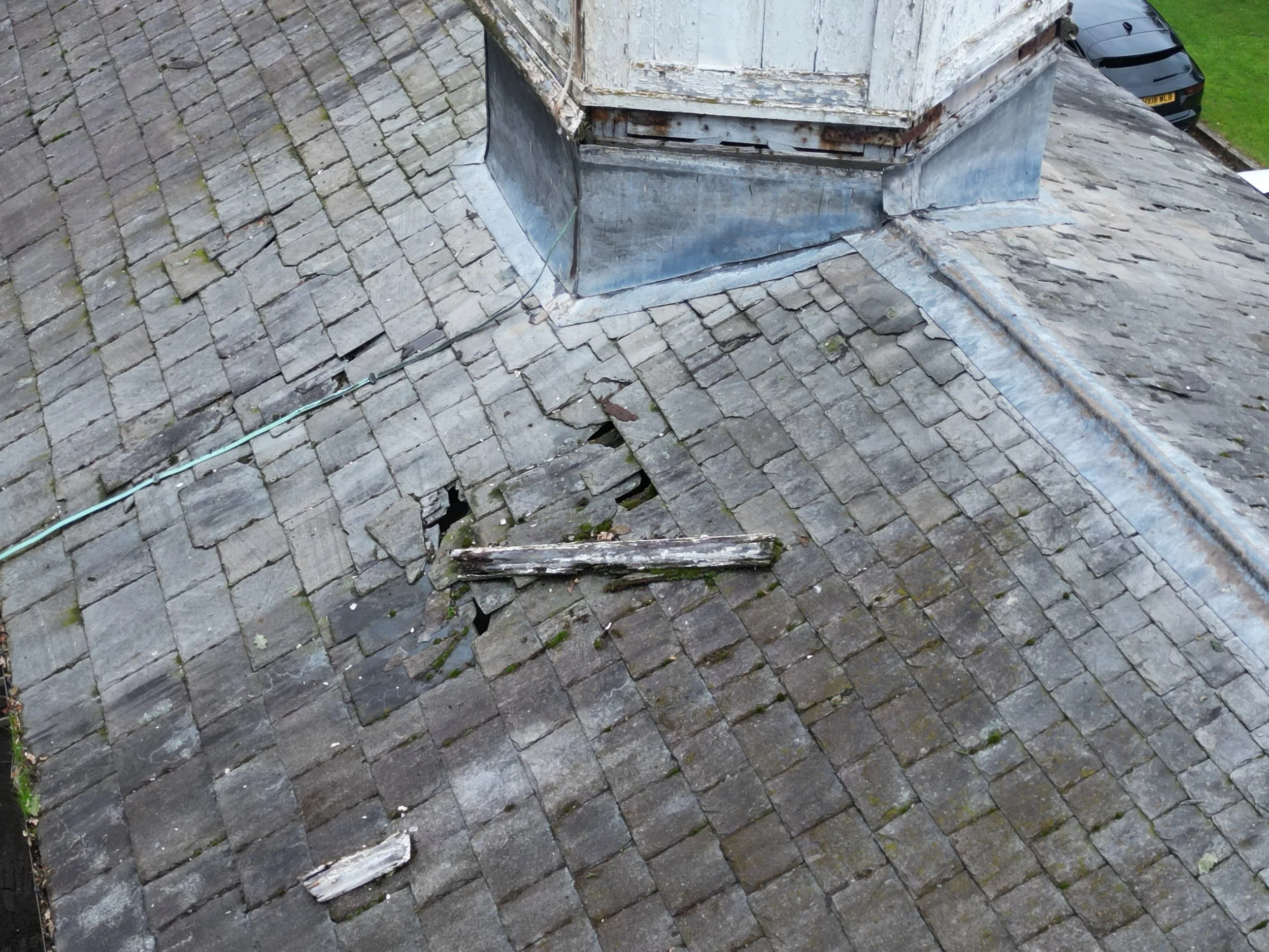 A rooftop with old, weathered gray shingles, a metal chimney flashing, and visible damage in some shingles. A black car is parked on a driveway and a grassy area is visible in the background.