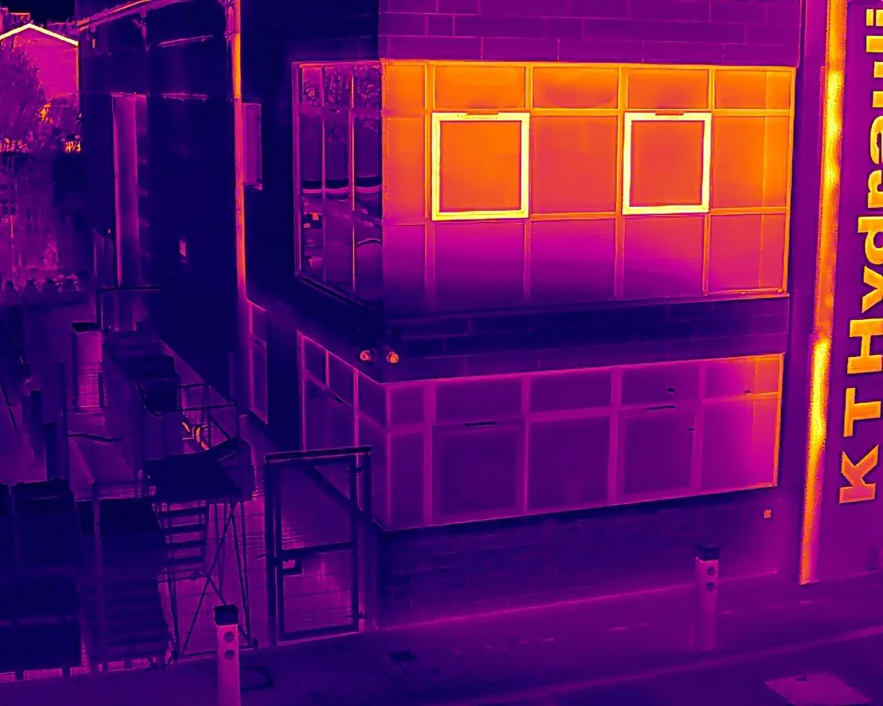 Infrared image of a building's exterior showing a corner with a large sign on the right side. The building has windows and a balcony, with colors indicating heat levels varying from purple to yellow.
