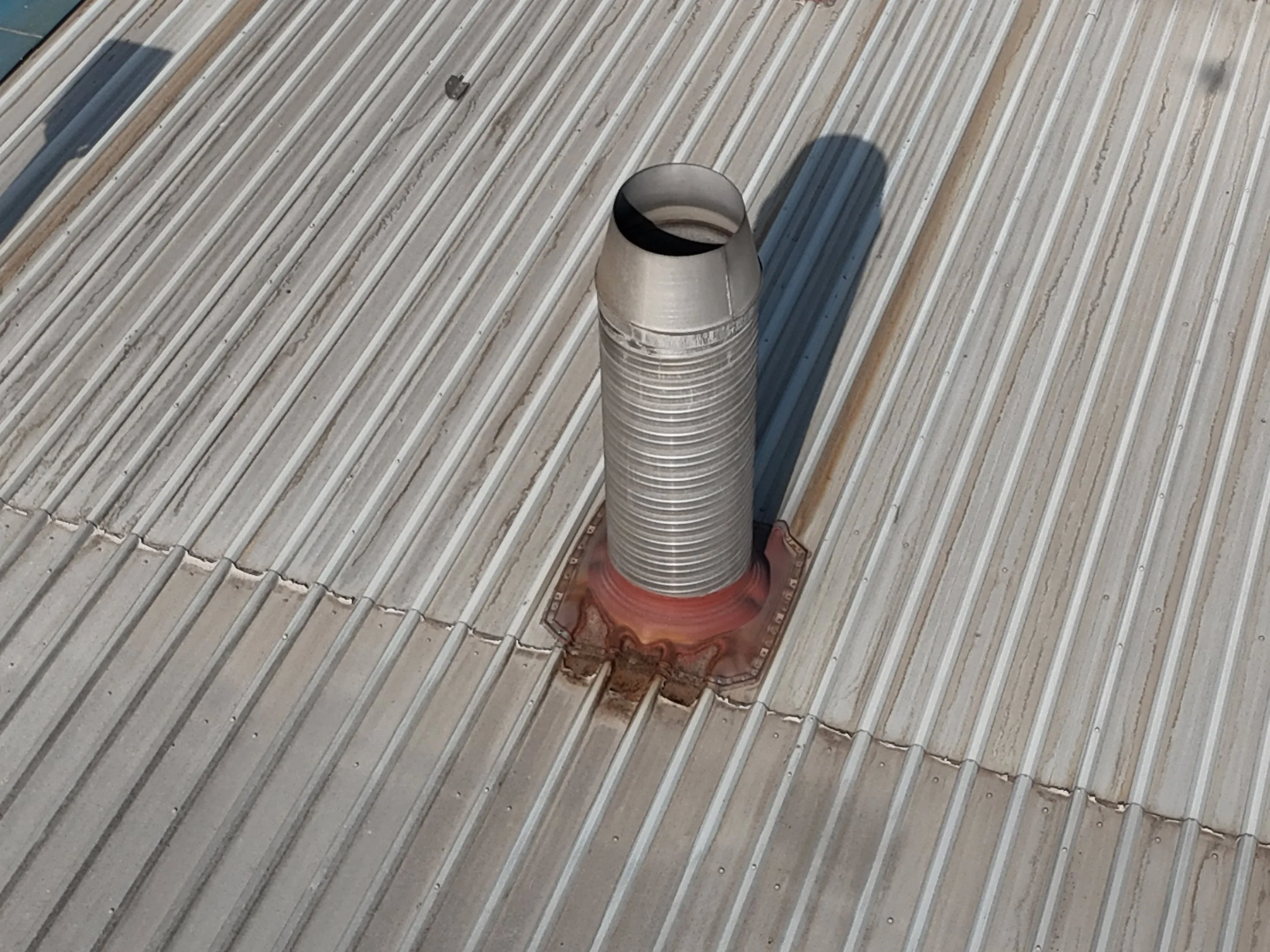 A metallic vent pipe protrudes from a metal rooftop with parallel ridges, casting a shadow.