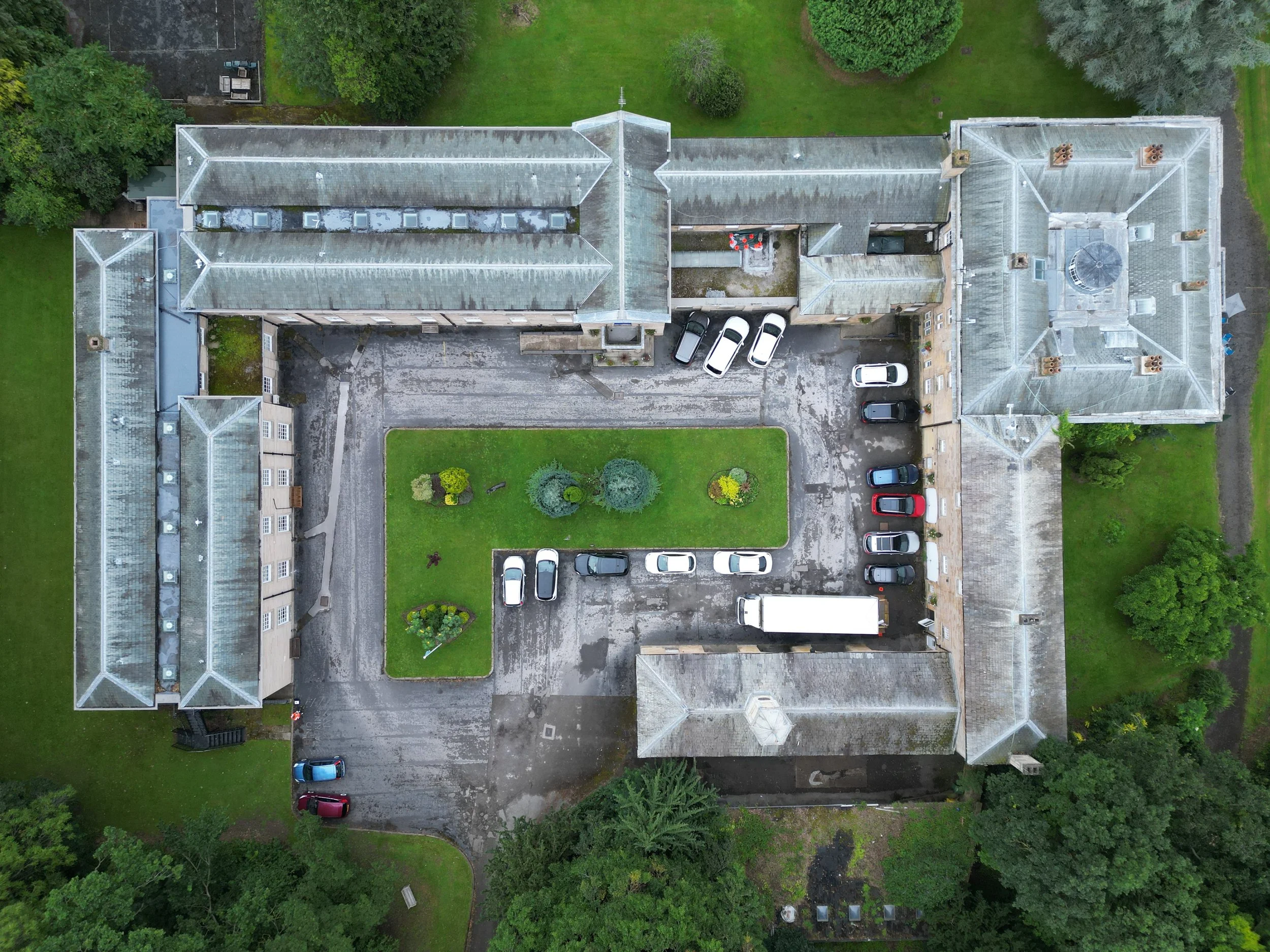 An aerial view of a large, multi-section building with a gray, weathered roof, green lawn, trees, and parked cars.