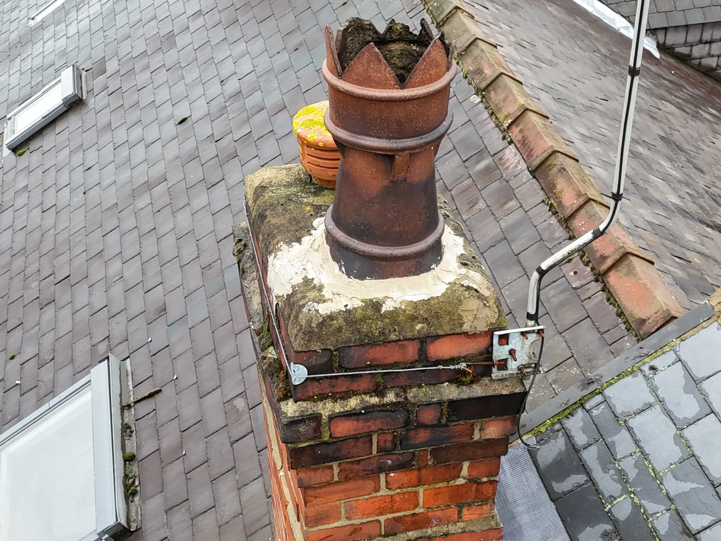 A brick chimney with a metal chimney cap on a sloped roof, with moss and some debris around it, and nearby roof tiles.