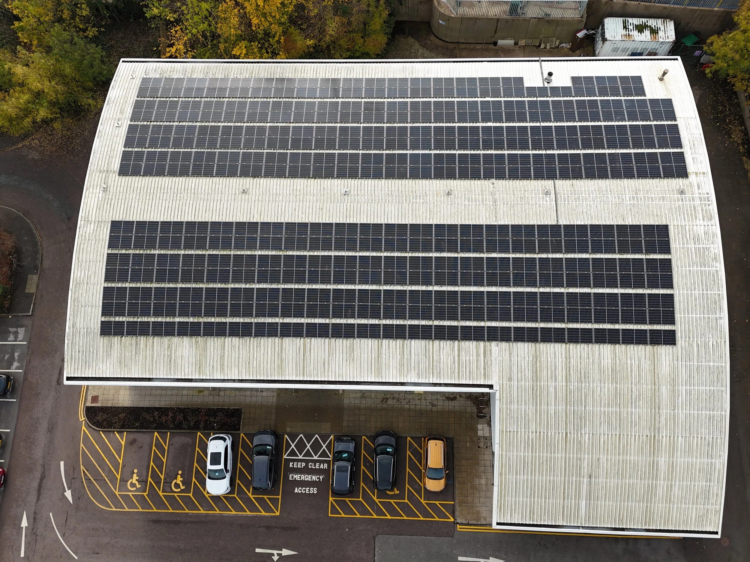 Top-down view of a building with solar panels on its metal roof, adjacent to a parking lot with designated accessible spaces and a sign that reads 'Keep Clear Emergency Access'.