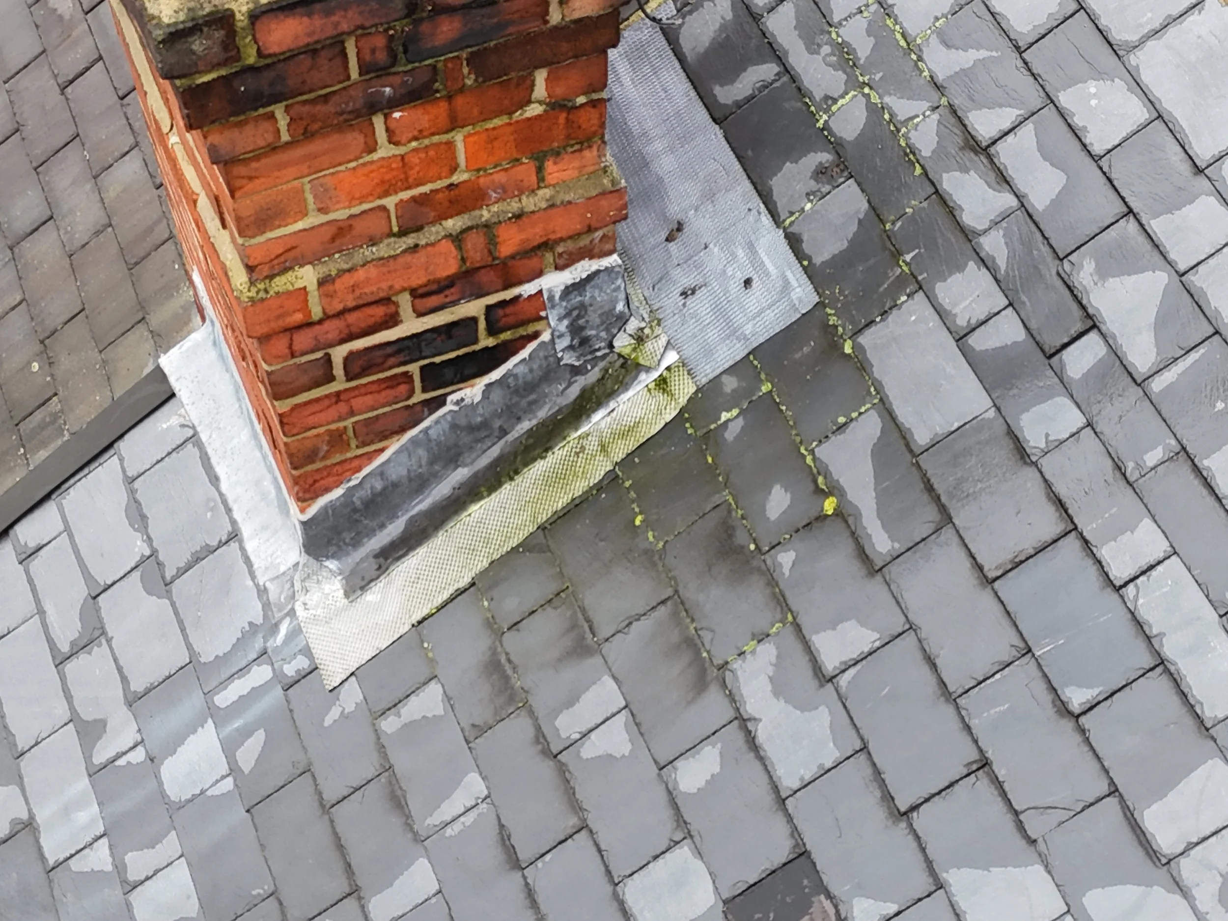 Close-up of a brick chimney with a metal flashing at its base, and cobblestone paving on the ground surrounding it. The mortar on the chimney is visible, and there are some small patches of moss or dirt.