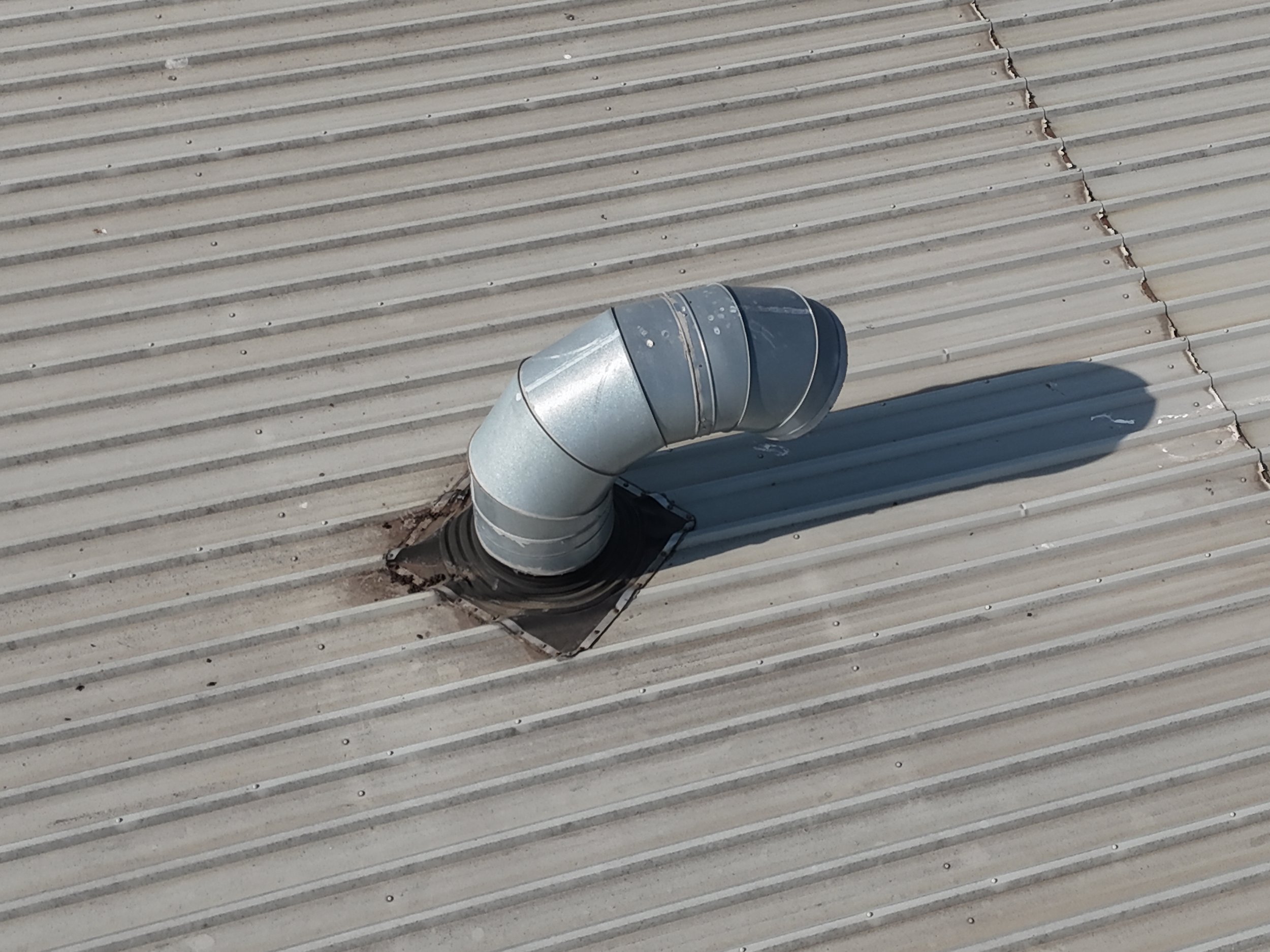 A metal ventilation pipe on a corrugated metal roof with visible seams and a shadow cast across the surface.