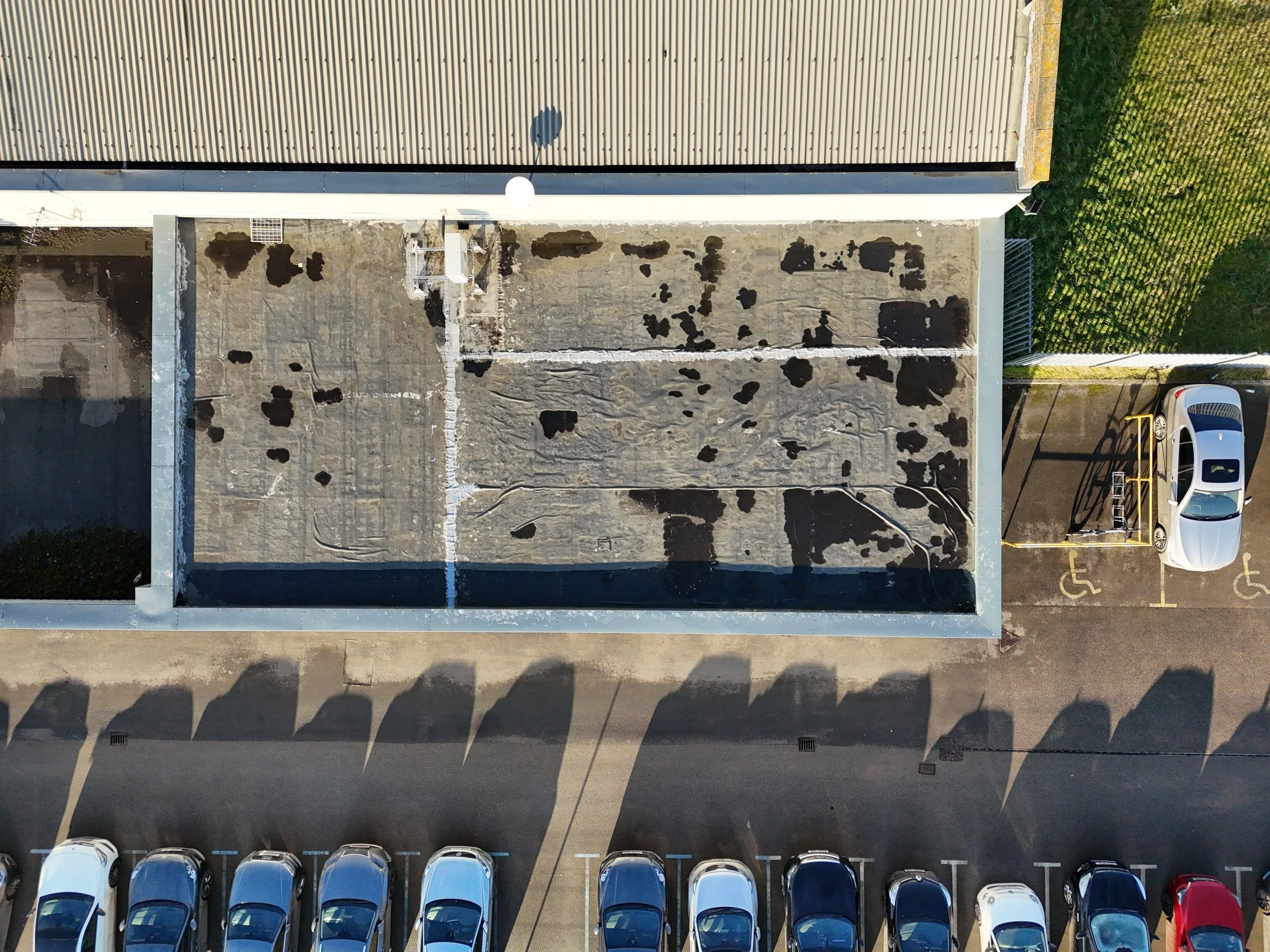 Aerial view of a parking lot with cars, an area of the roof with dark stains and a crack, and an adjacent grassy area.
