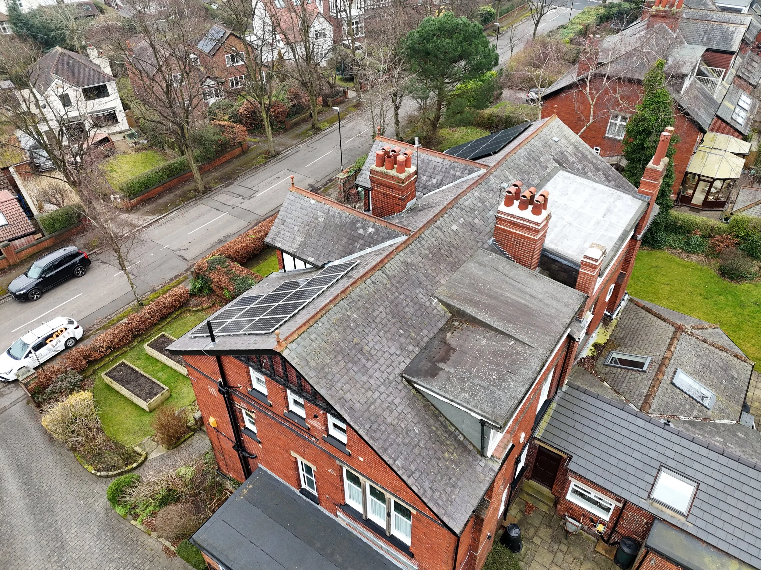 Aerial view of a residential neighborhood with brick houses, parked cars along the street, and trees, some with leaves and some without, during an overcast day.