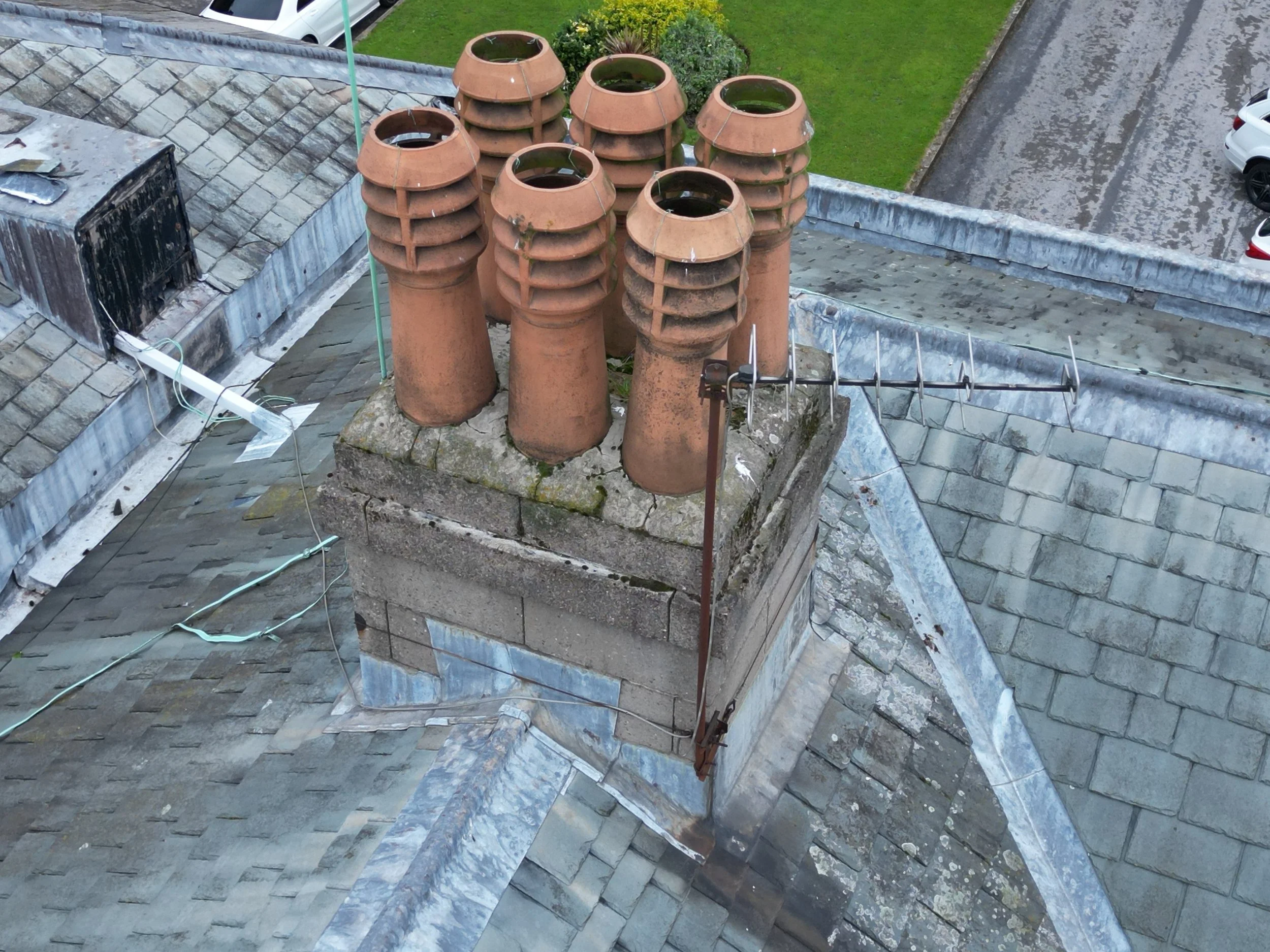 Rooftop with multiple terracotta chimney pots, some filled with water, and a TV antenna. Nearby is a grassy area with parked white cars and a gravel road.