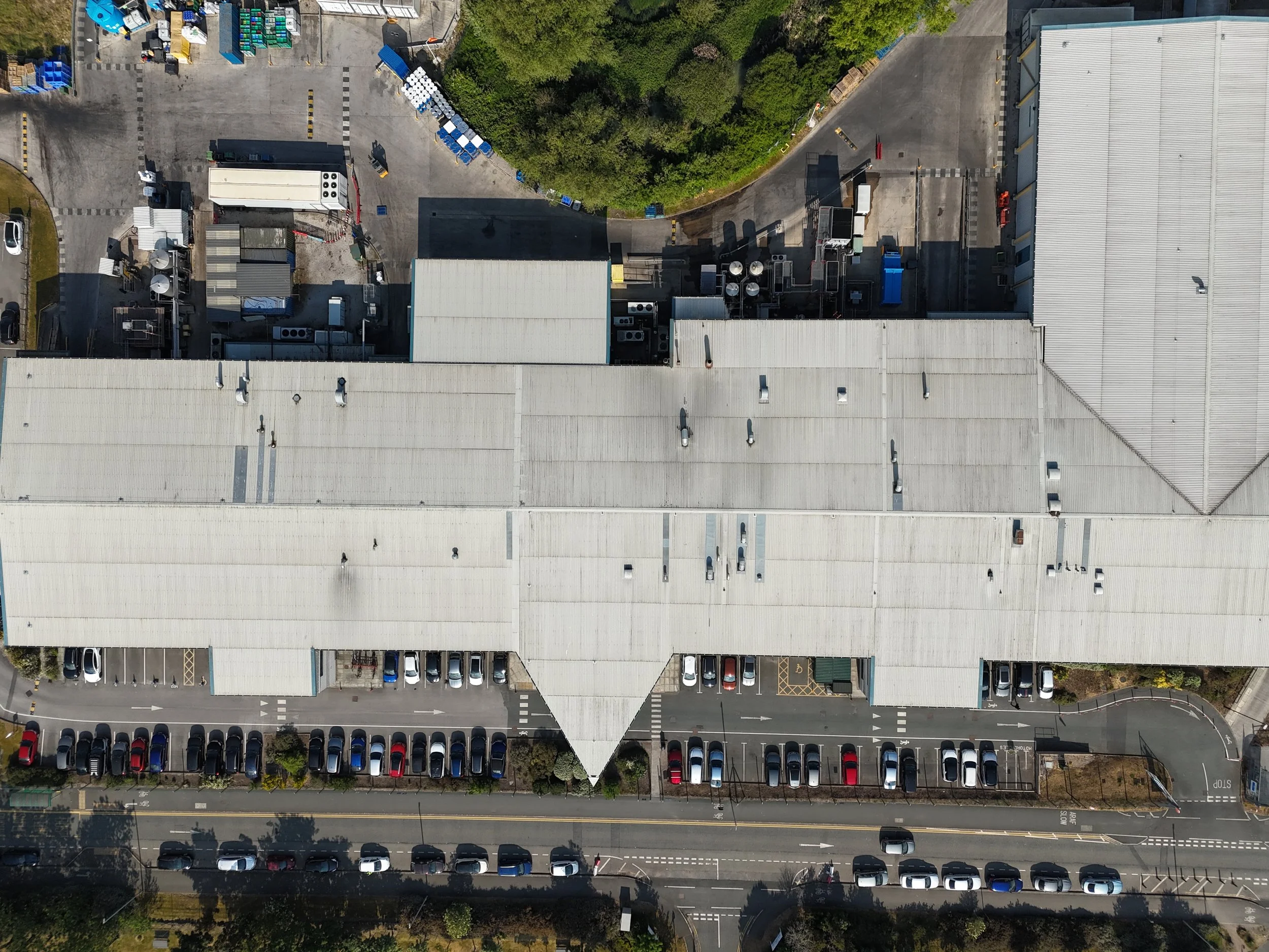 An aerial view of a large industrial or commercial building with a flat, light-colored roof. Surrounding the building are parking lots filled with many cars, and green areas with trees. There are loading areas, roads, and other smaller structures around the main building.