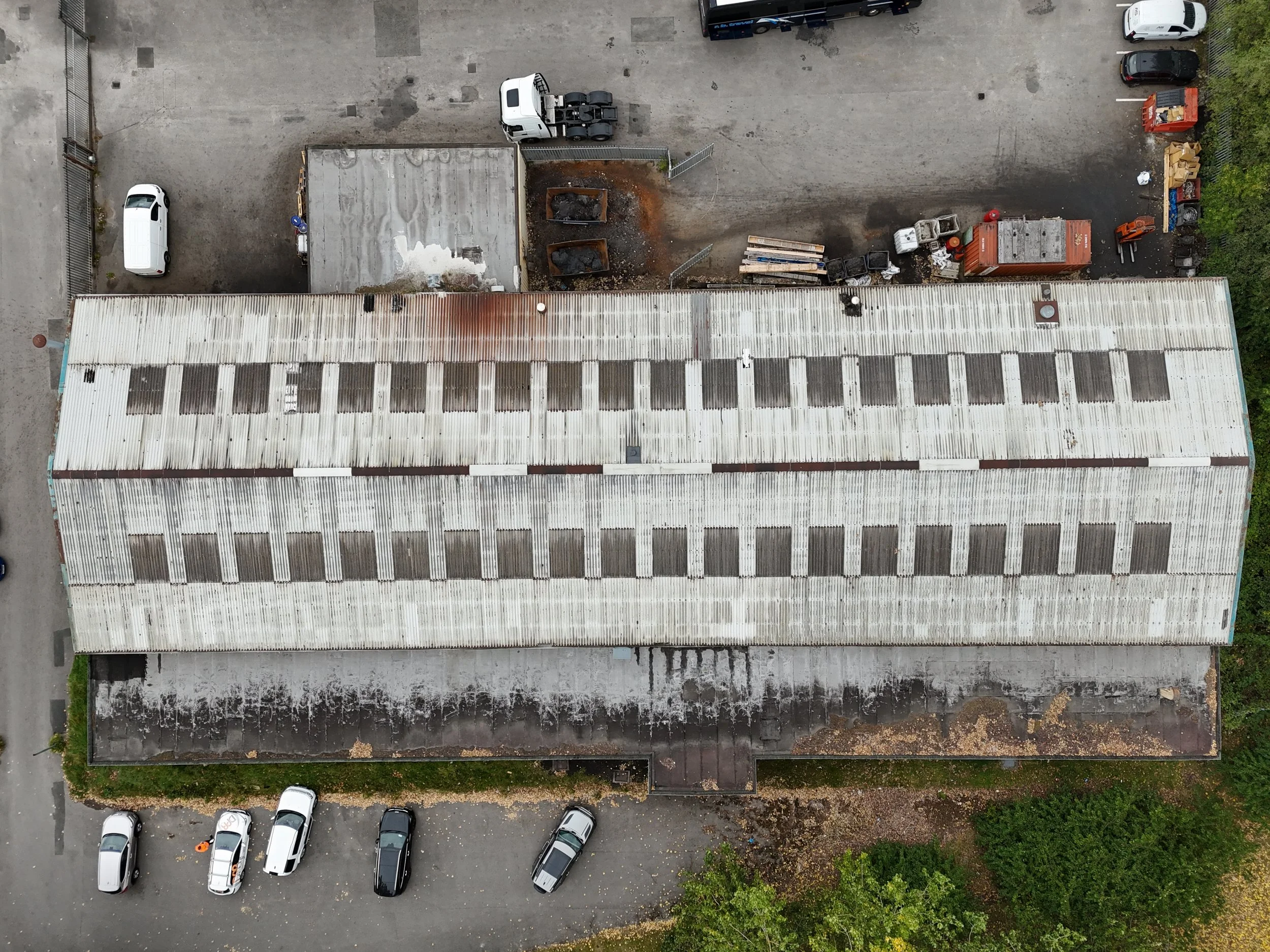 Aerial view of a parking lot with a large warehouse on the bottom half, featuring a white corrugated metal roof, and cars parked along the bottom edge. The upper part shows an open area with construction debris, gravel, and a white semi-truck. There are several other vehicles parked along the upper right and bottom edges of the image.