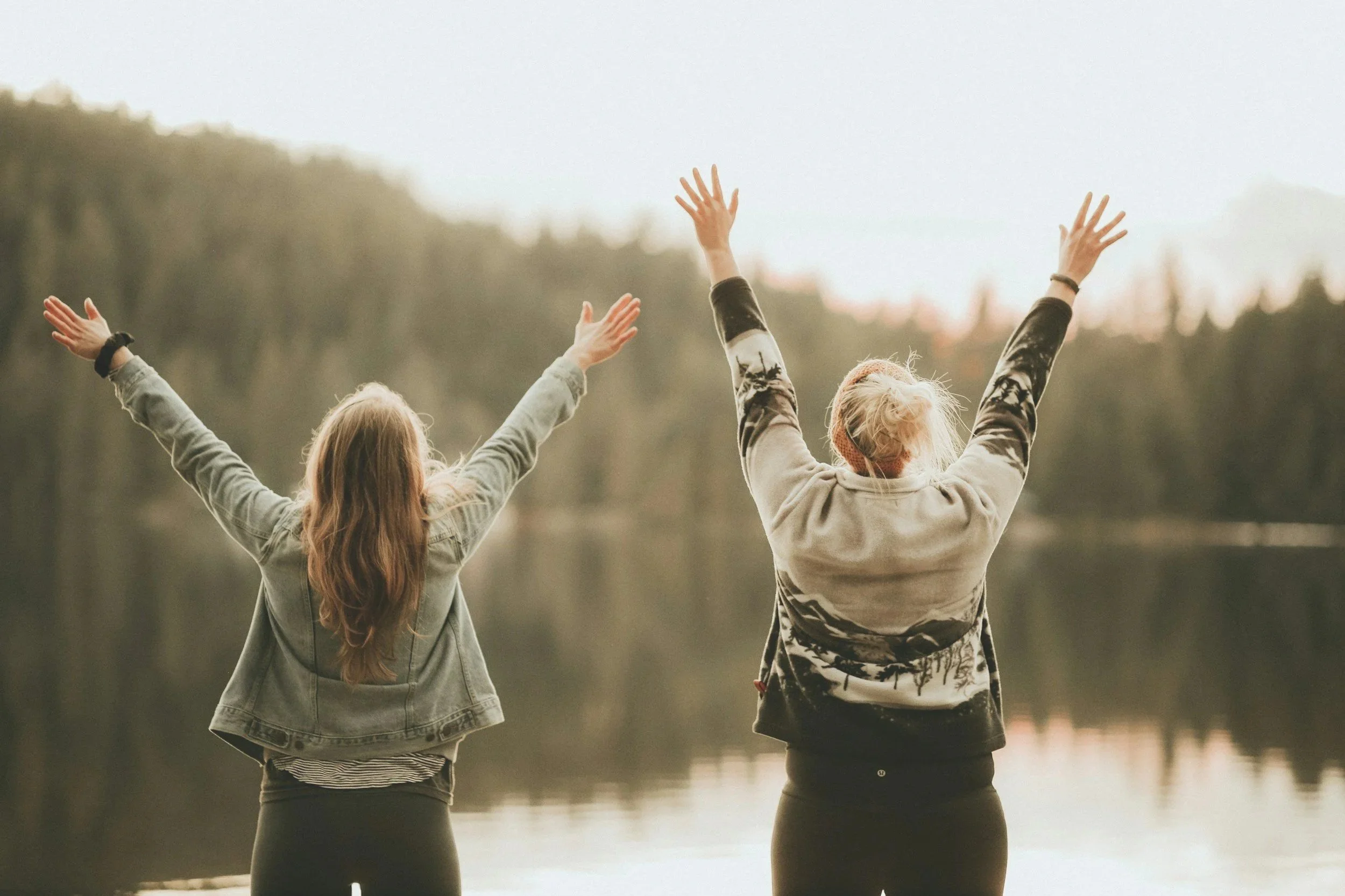 Two women with long hair, wearing casual jackets, with arms raised, standing outdoors near a calm body of water, during sunset or early evening.