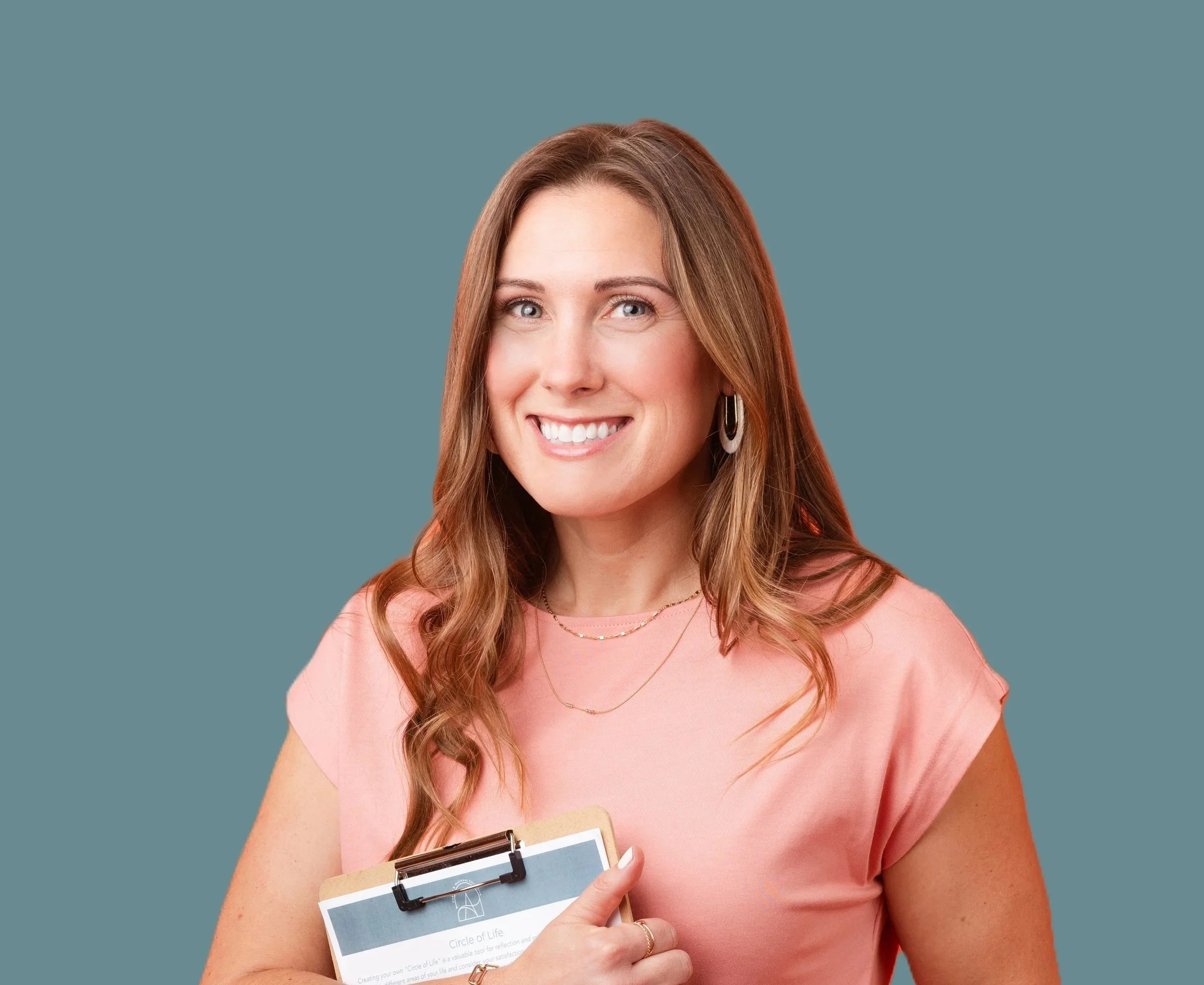 A woman with long, wavy auburn hair smiles while holding a clipboard and a booklet.