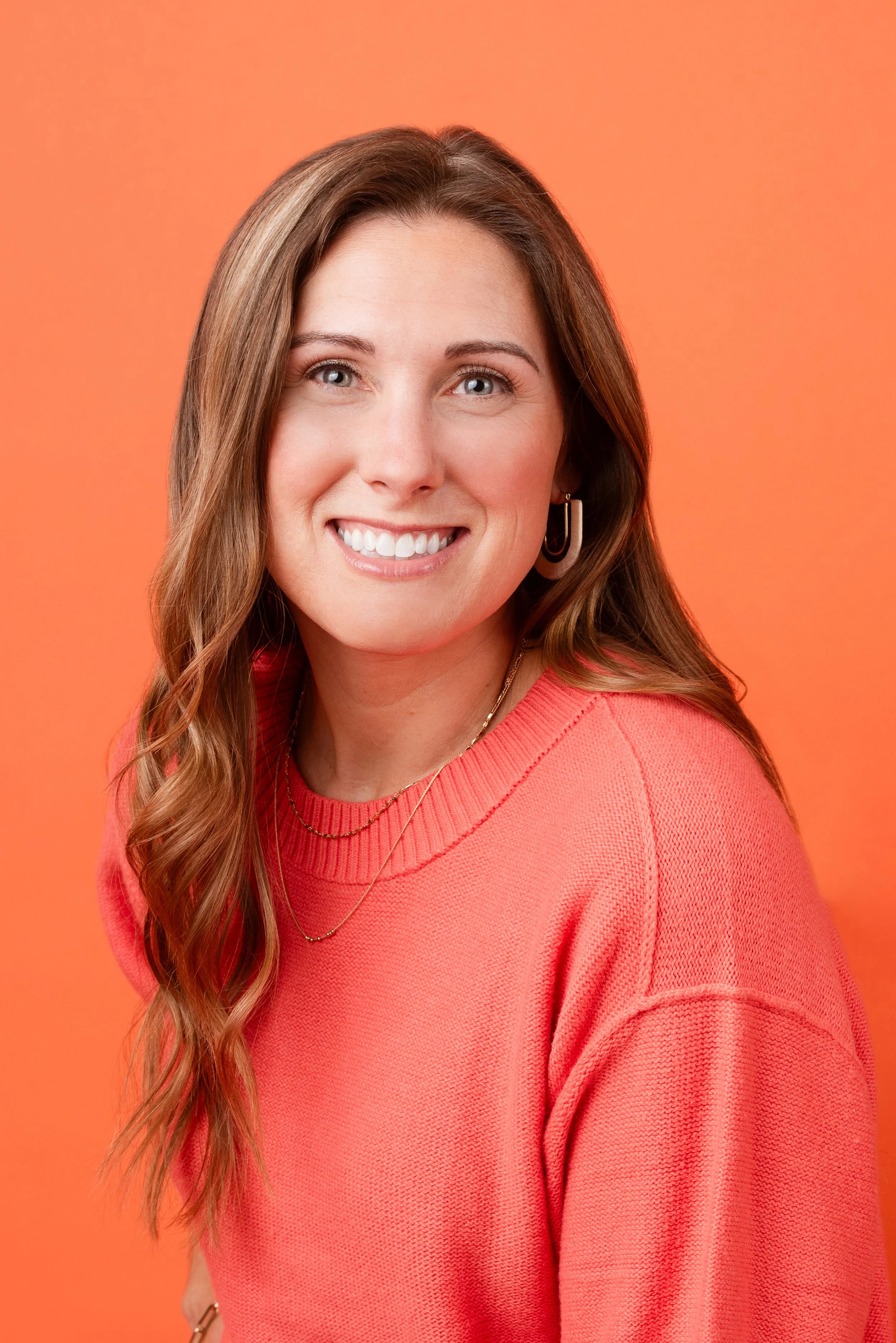 A young woman with long wavy brown hair, blue eyes, and a big smile wearing a coral knit sweater and gold jewelry against an orange background.