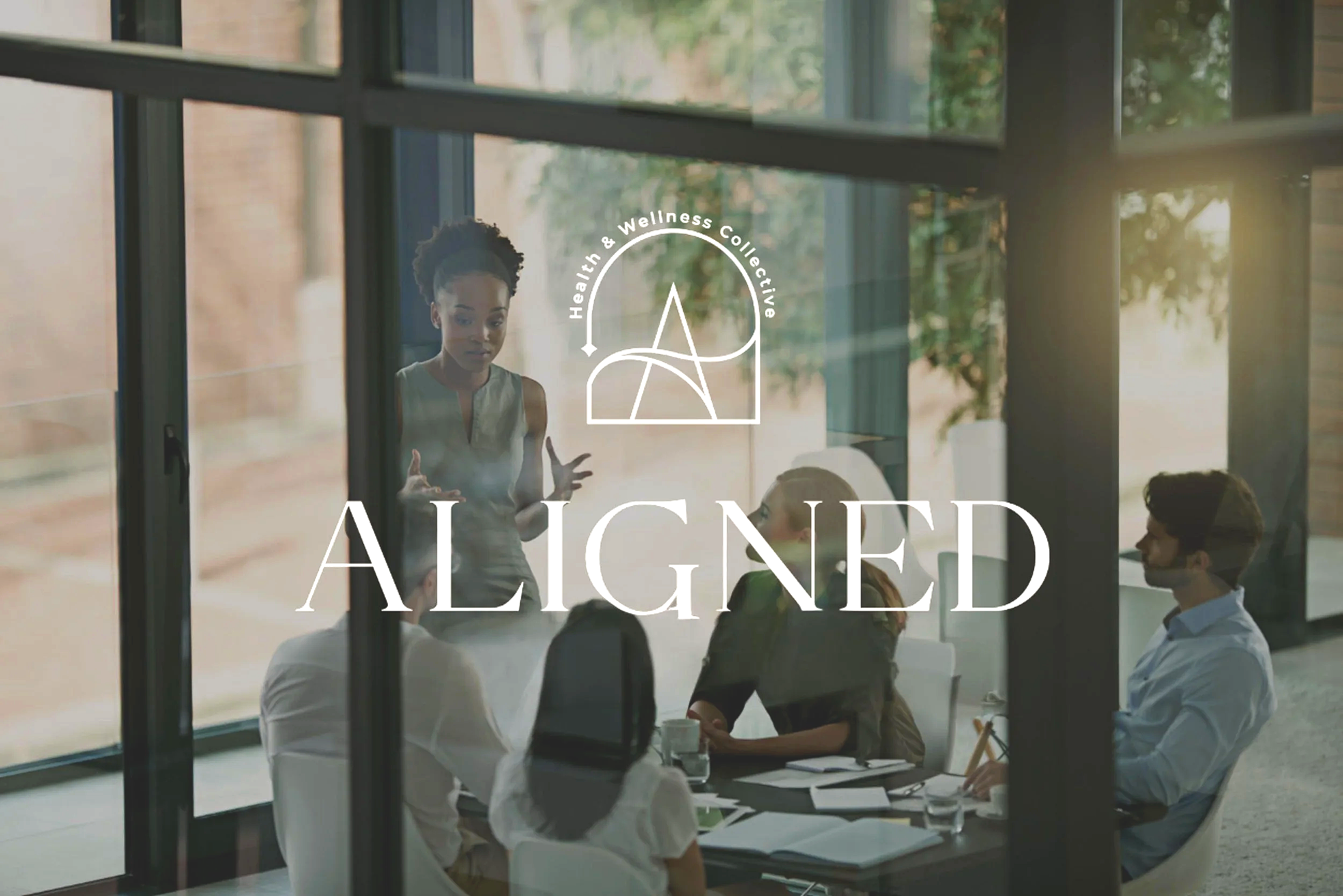 People meeting and discussing inside a modern office building with large glass windows, with the words 'Aligned' and a logo that reads 'Health & Wellness Collective' on the window.