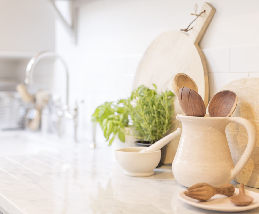 A clean kitchen countertop with wooden utensils in a ceramic pitcher, a bowl with a pestle, and a potted basil plant. Cutting boards are leaning against the wall.