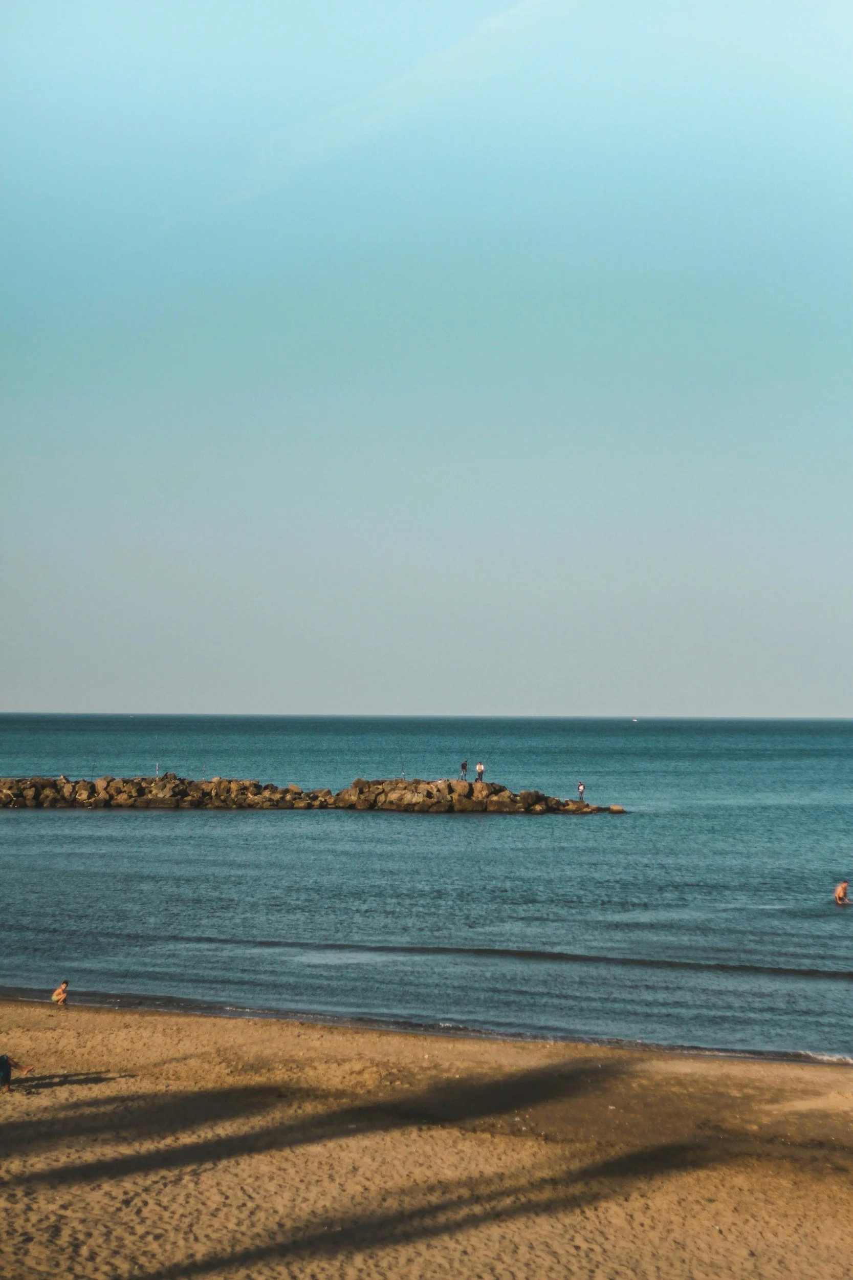 A calm beach scene with a sandy shore at the bottom, a breakwater extending into the ocean with people standing on it, and the vast ocean with gentle waves under a clear blue sky.