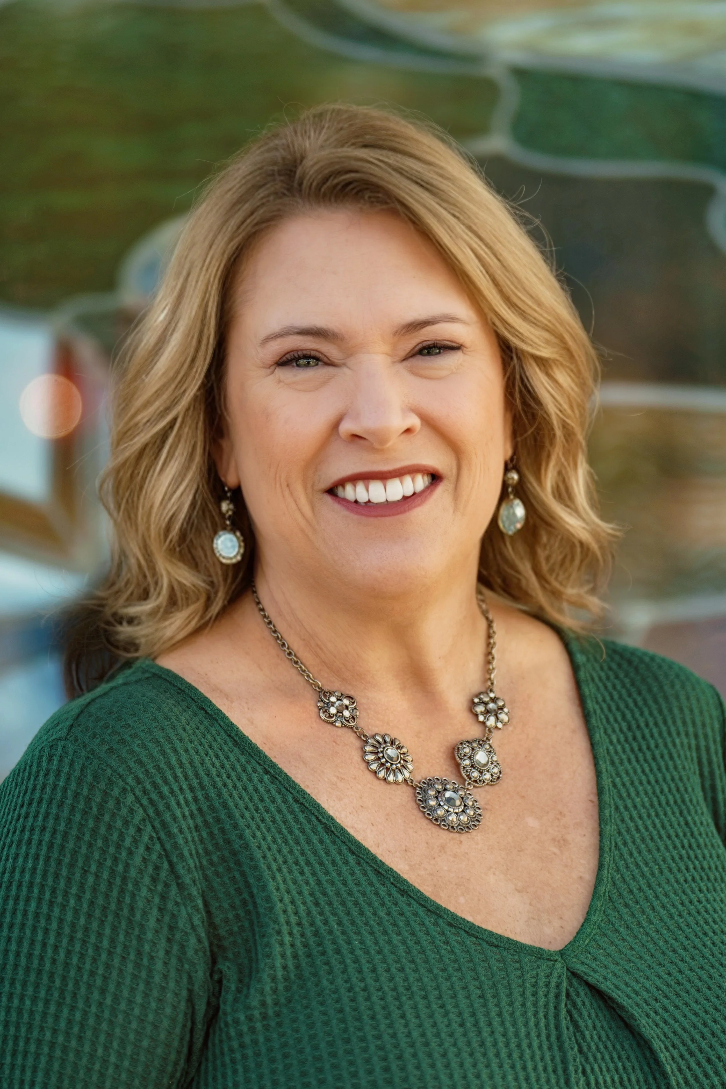 A woman smiling with blonde hair, wearing jewelry and a green textured top, standing outdoors with blurred colorful background.