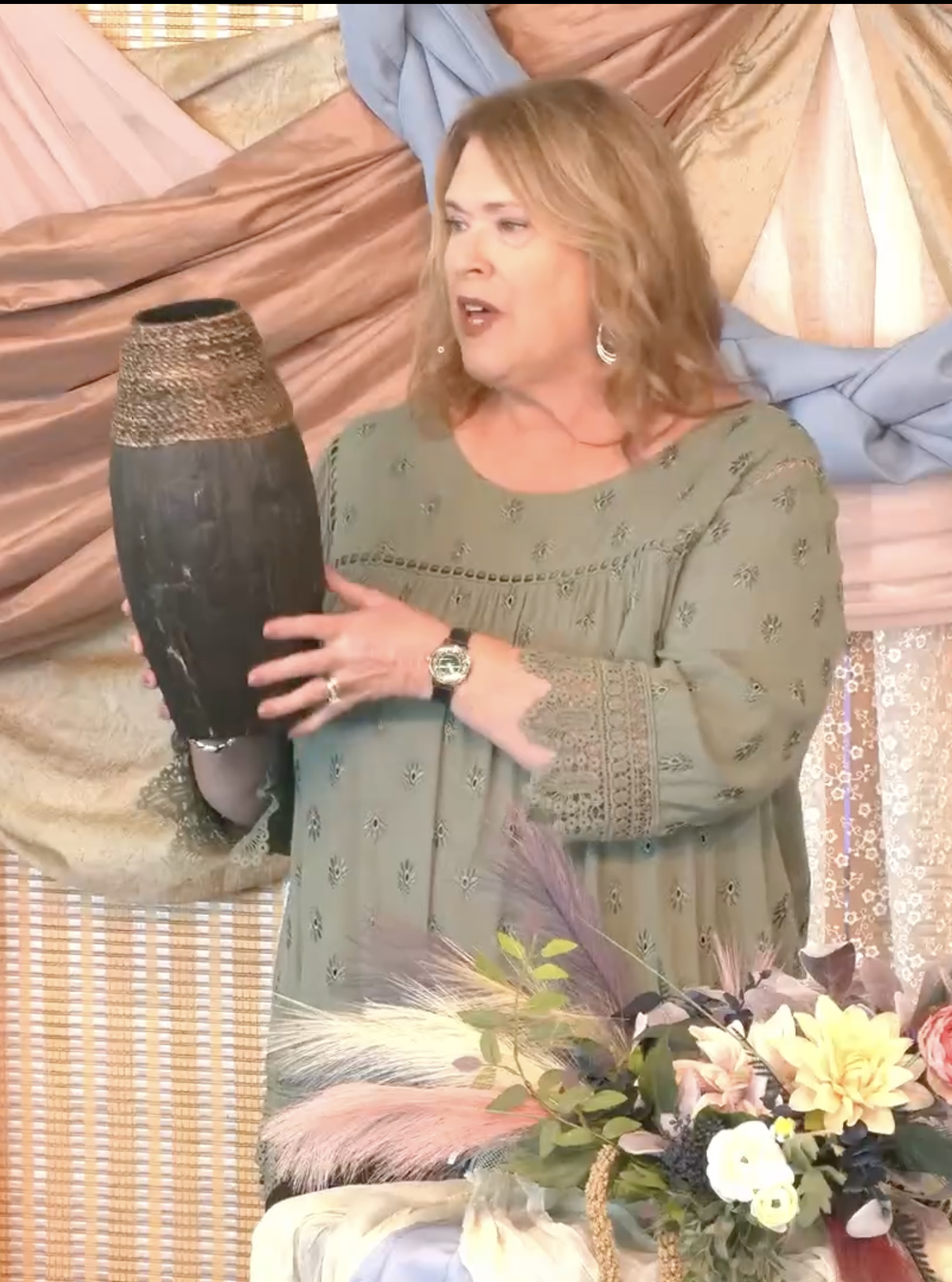 A woman with shoulder-length blonde hair, wearing a green dress with embroidered details, holds a dark vase. She is standing in front of a background decorated with pink, blue, and peach draped fabric and a floral arrangement with yellow, pink, and purple flowers.