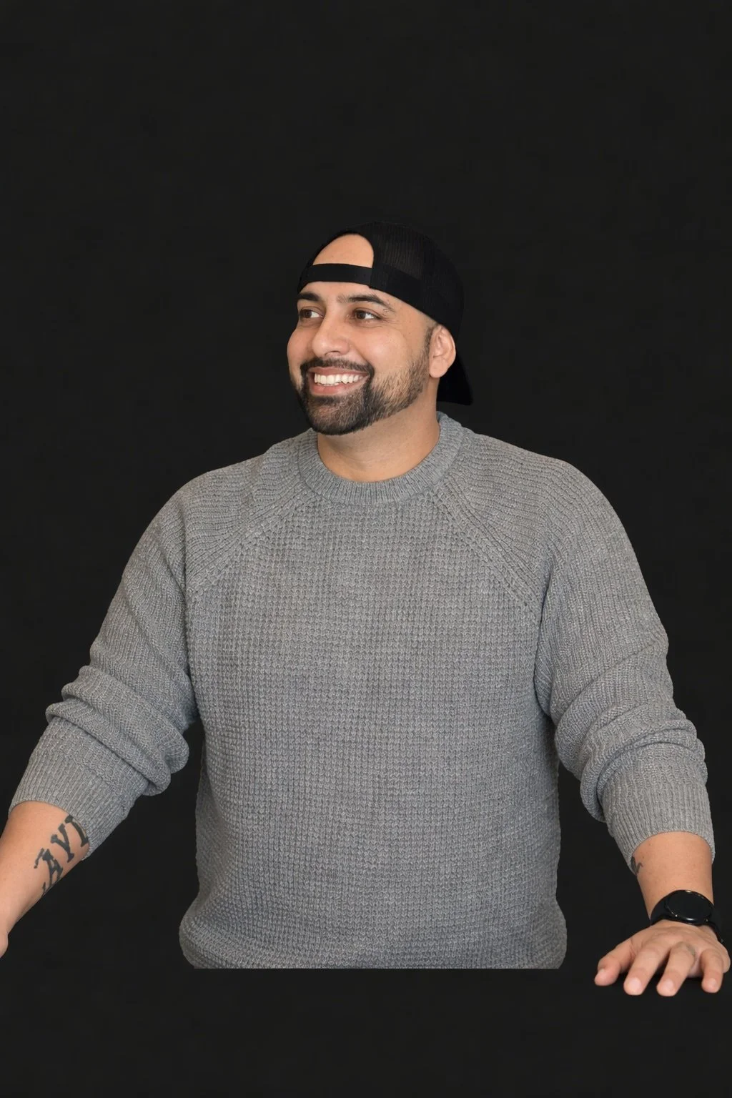 A man smiling, wearing a gray sweater and a black baseball cap backward, standing in front of a black background.