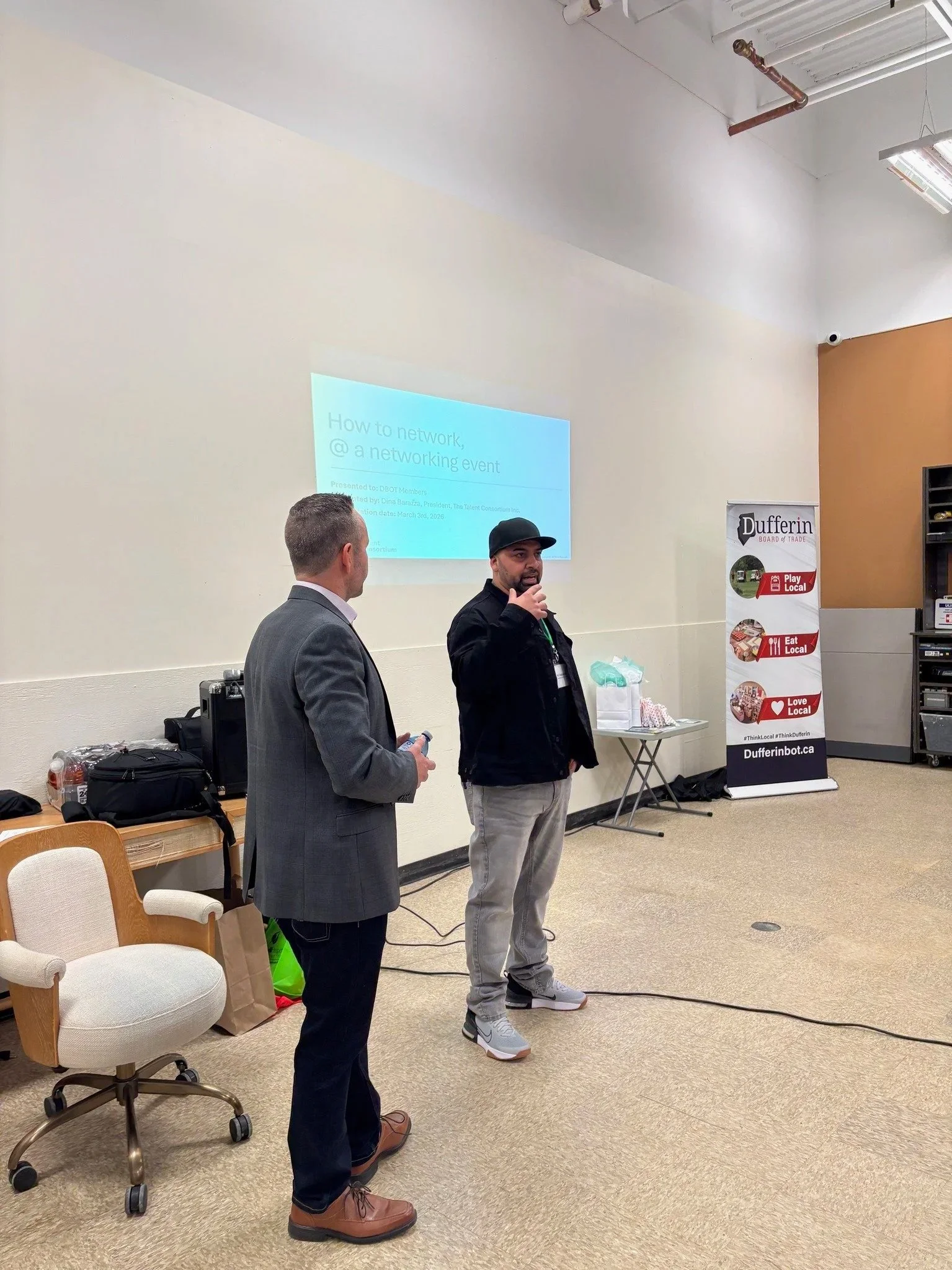 Two men are standing in front of a white wall, with one speaking into a microphone. A presentation slide titled 'How to network @ a networking event' is projected on the wall. There is a table with supplies and a banner that reads 'Dufferin' with slogans about supporting local businesses.
