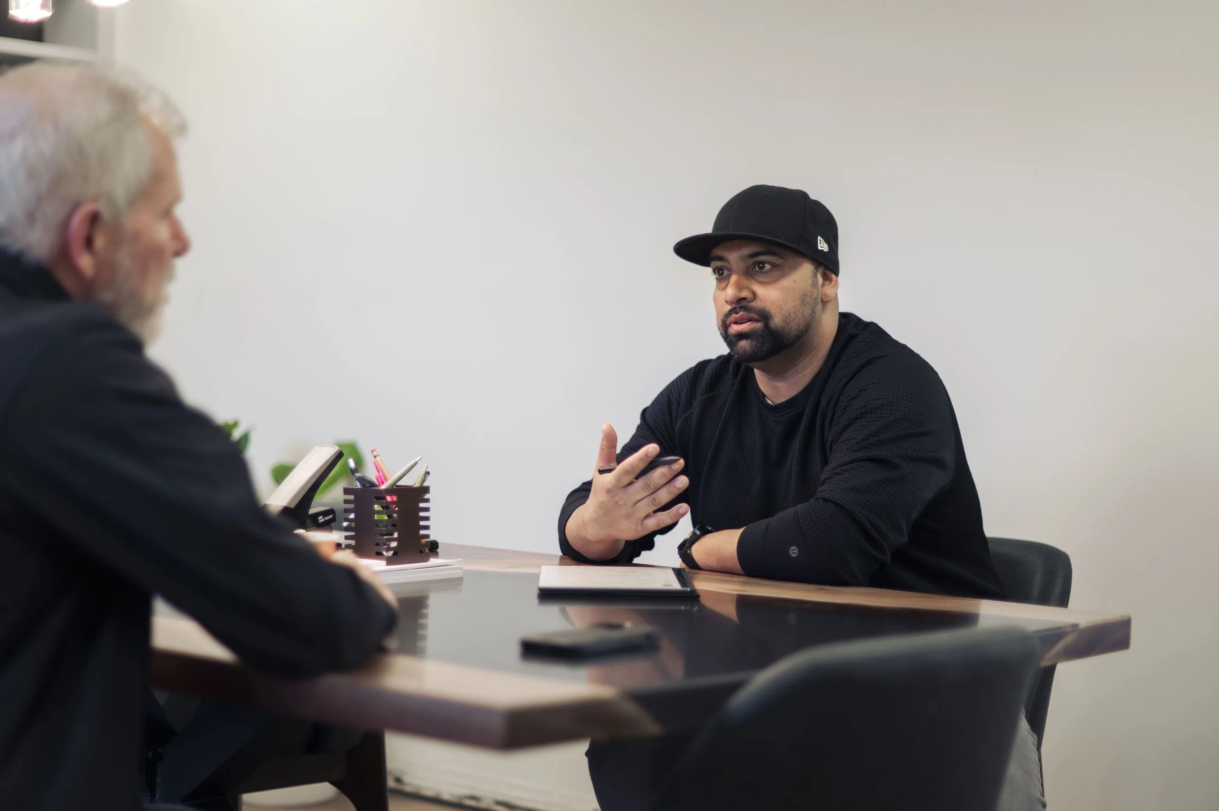 Two men having a discussion across a desk in an office, one man with a beard and gray hair, the other man wearing a black cap and black shirt, with notebooks and stationery on the desk.