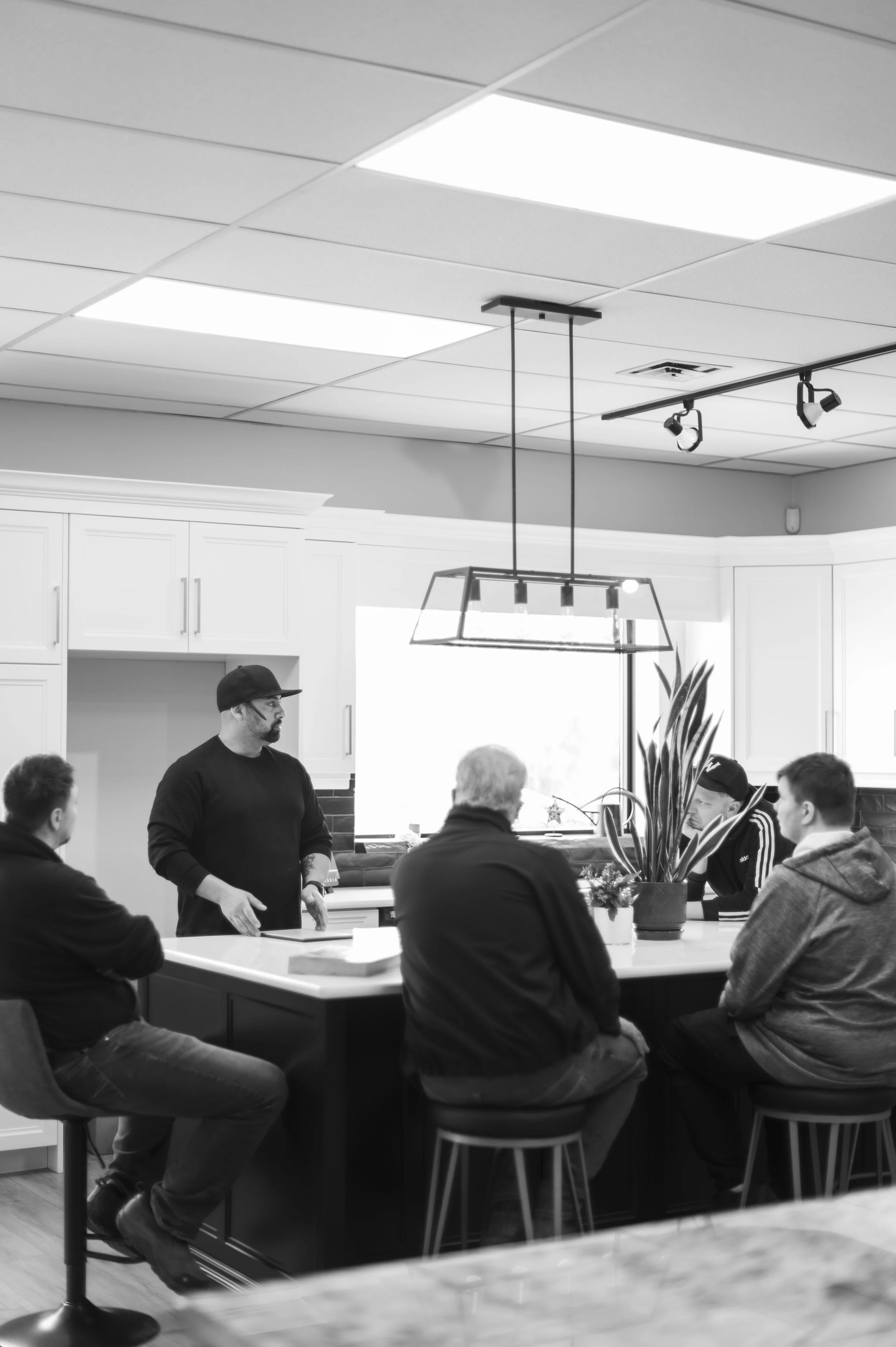 A group of five men sitting around a kitchen island, with one man standing and talking, in a modern kitchen with white cabinets and pendant lighting.
