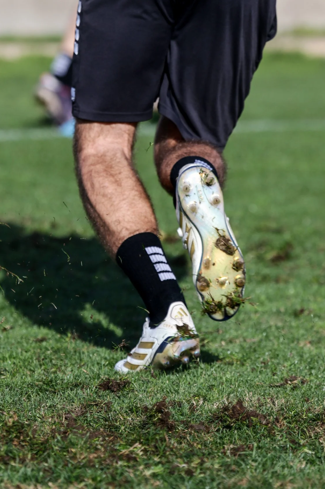 Fußballspieler beim Sprint auf einem Rasenplatz, mit rausfederndem Bein, Grasspritzer in der Luft, trägt schwarze Shorts, weiße Fußballschuhe und schwarze Socken.