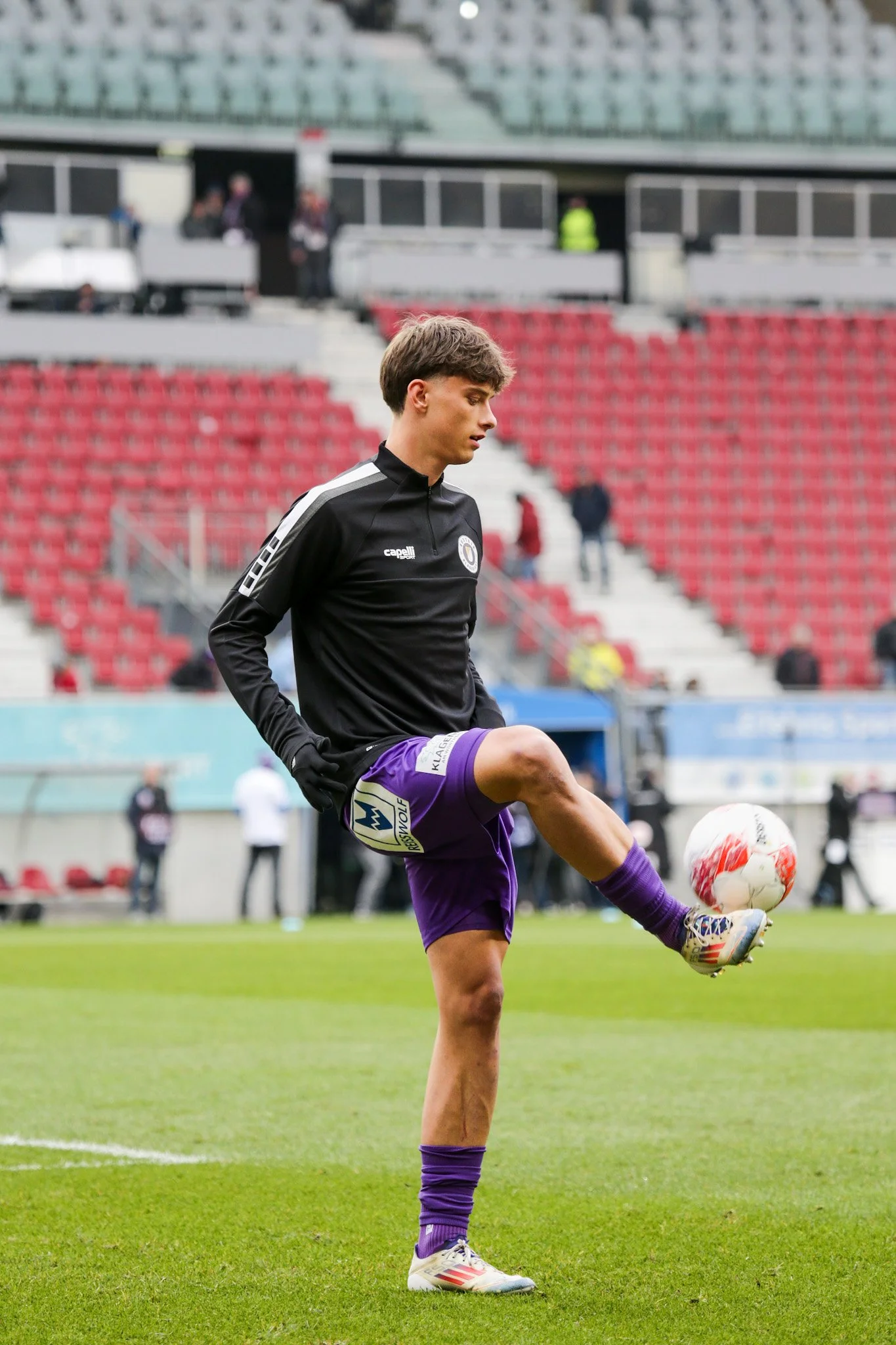 Ein junger Fußballspieler in schwarzem Trikot und lila Shorts beim Aufwärmen auf dem Spielfeld, der den Ball mit dem Fuß in der Luft kontrolliert.