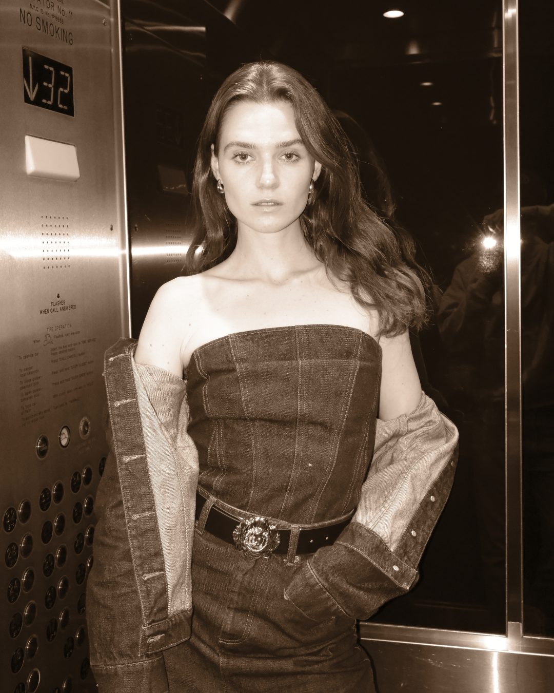 Junge Frau mit langem Haar in einem gestreiften, ärmellosen Top und einer Jeansjacke, die von den Schultern rutscht, steht in einem Aufzug und schaut in die Kamera.