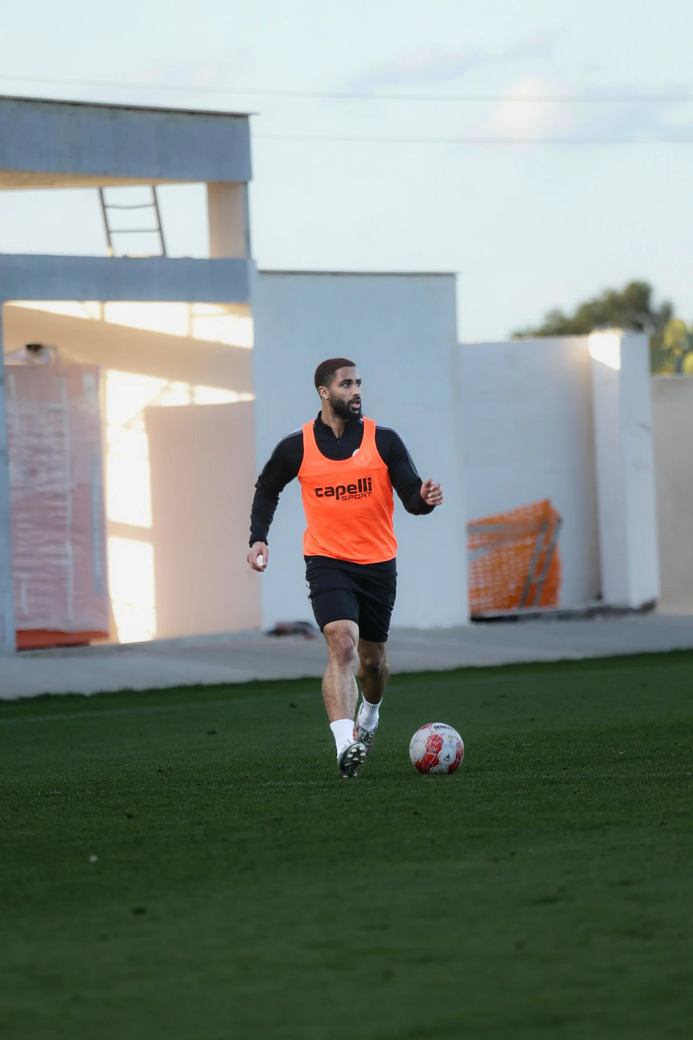 Ein Mann spielt Fußball auf einem Rasenplatz, trägt ein orangefarbenes Trainingsweste mit dem Schriftzug 'Capelli Sport'.