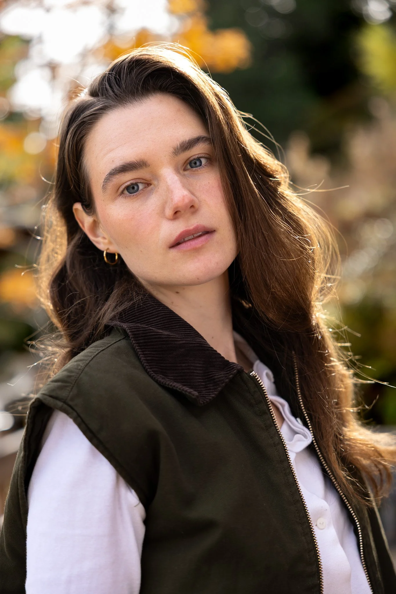 Porträt einer jungen Frau mit braunen Haaren vor einem herbstlichen Hintergrund, trägt eine weiße Bluse und eine dunkle Jacke.