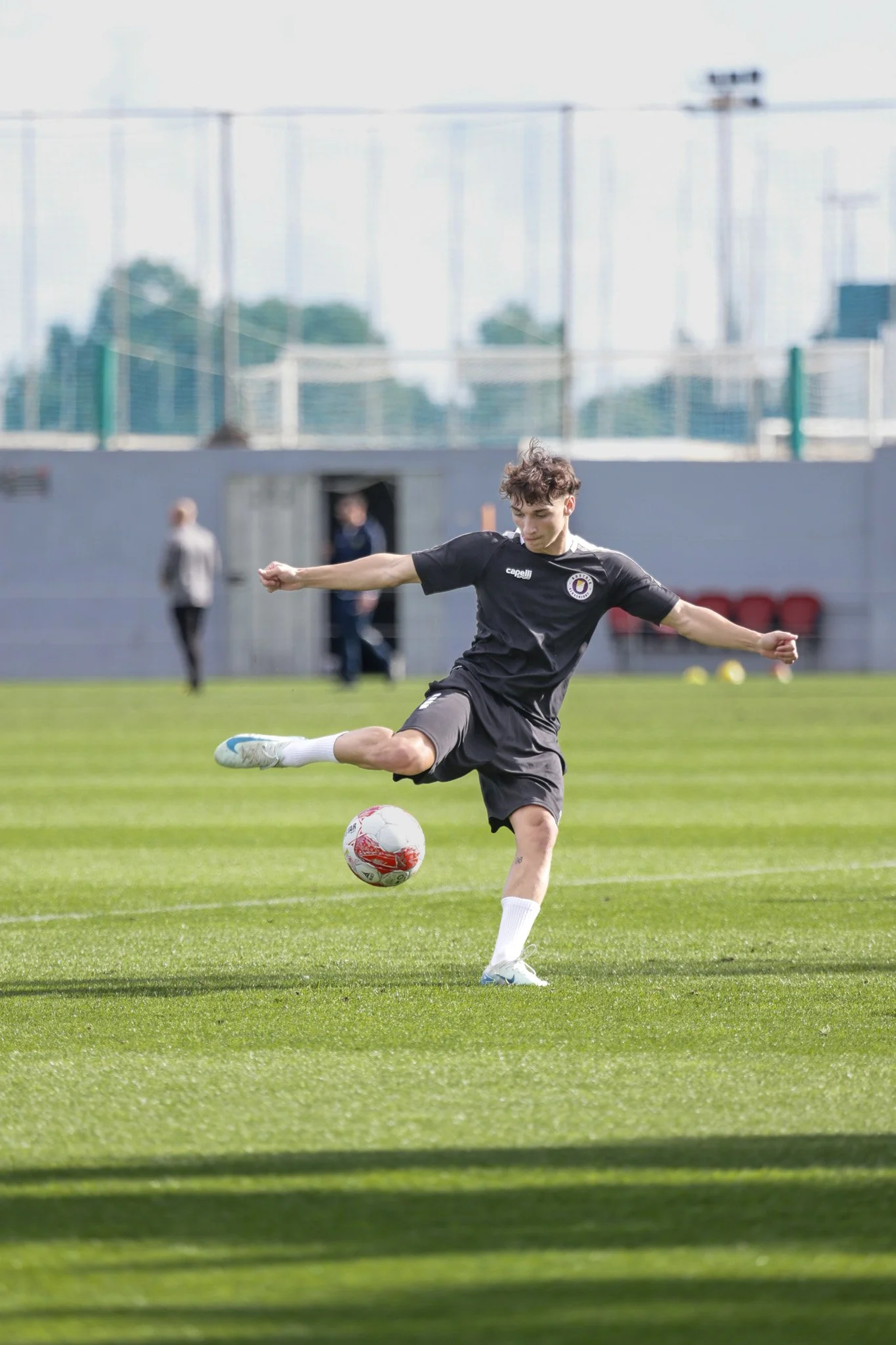 Junge Fußballspieler trainiert auf einem grünen Fußballfeld, springt, um den Ball zu kicken, während er in einem schwarzen Trikot steht.