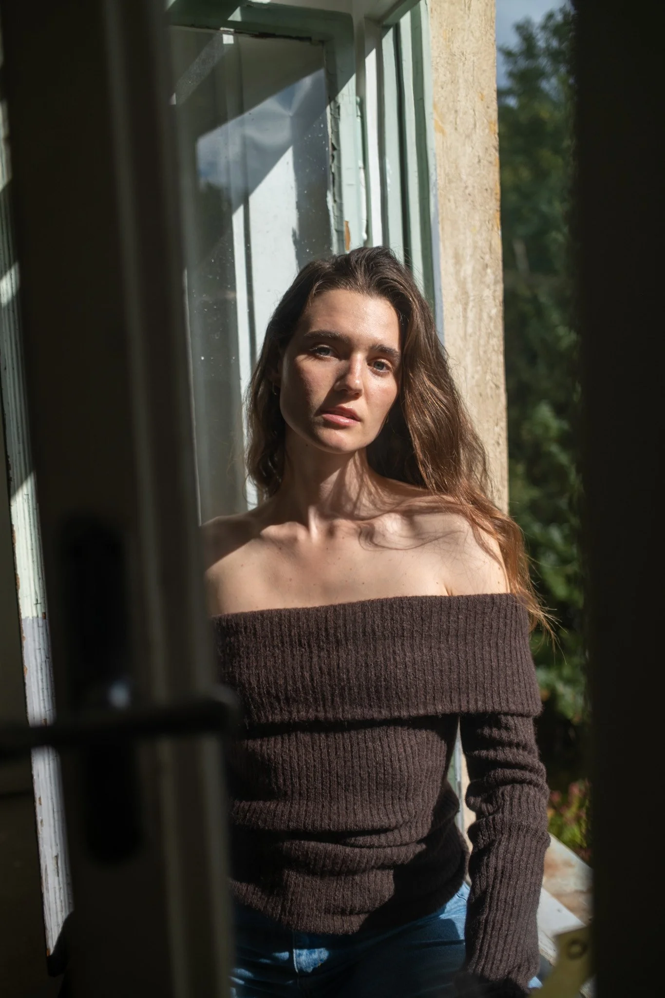 Junge Frau mit braunem Pullover sitzt an einem Fenster, Sonnenlicht fällt auf ihr Gesicht, draußen sind Bäume sichtbar.