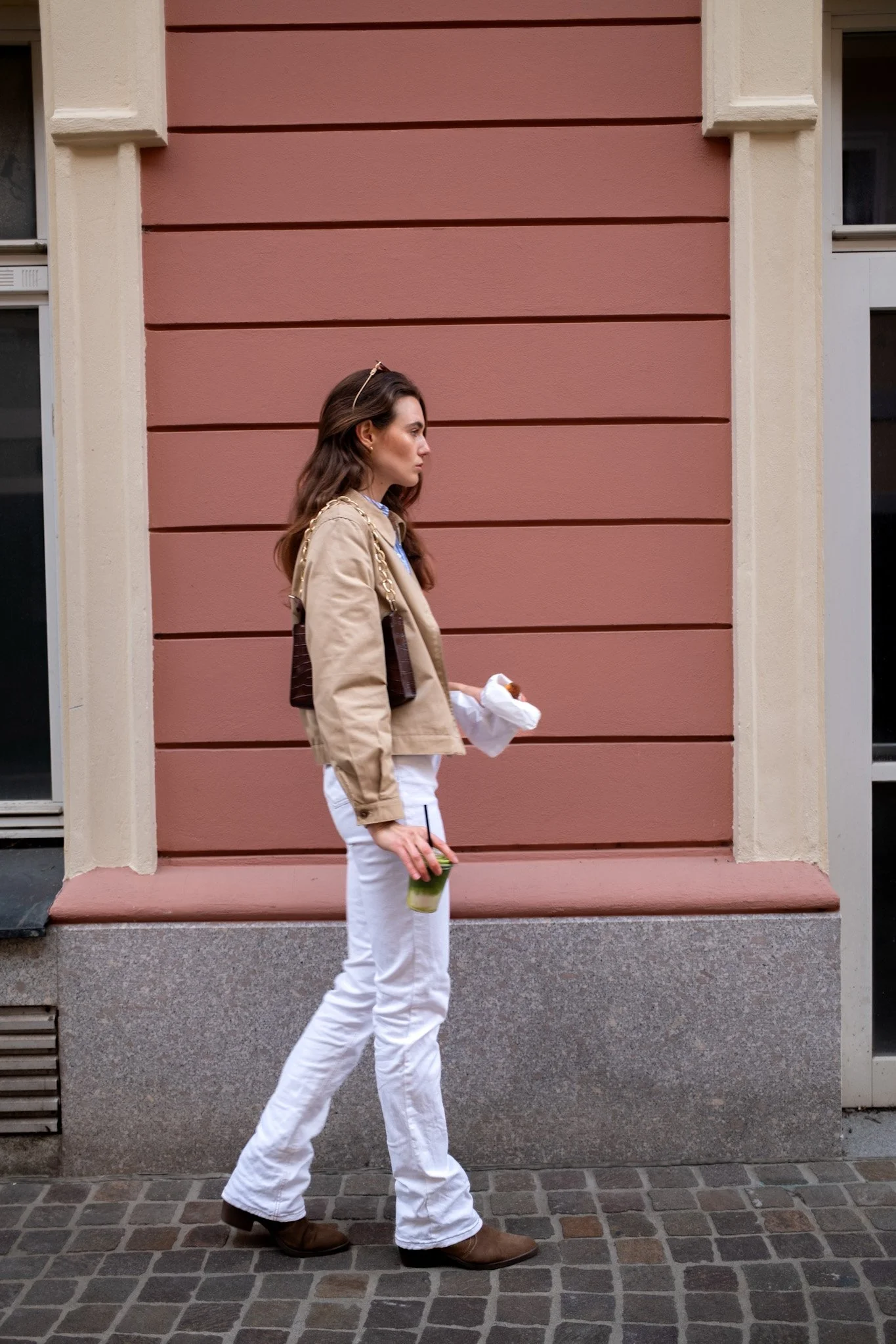 Frau geht auf einem Bürgersteig neben einer rosa Wand, trägt eine beige Jacke, weiße Hosen, braune Schuhe, hält eine grüne Smoothie und ein Stück Pizza in Papierverpackung.