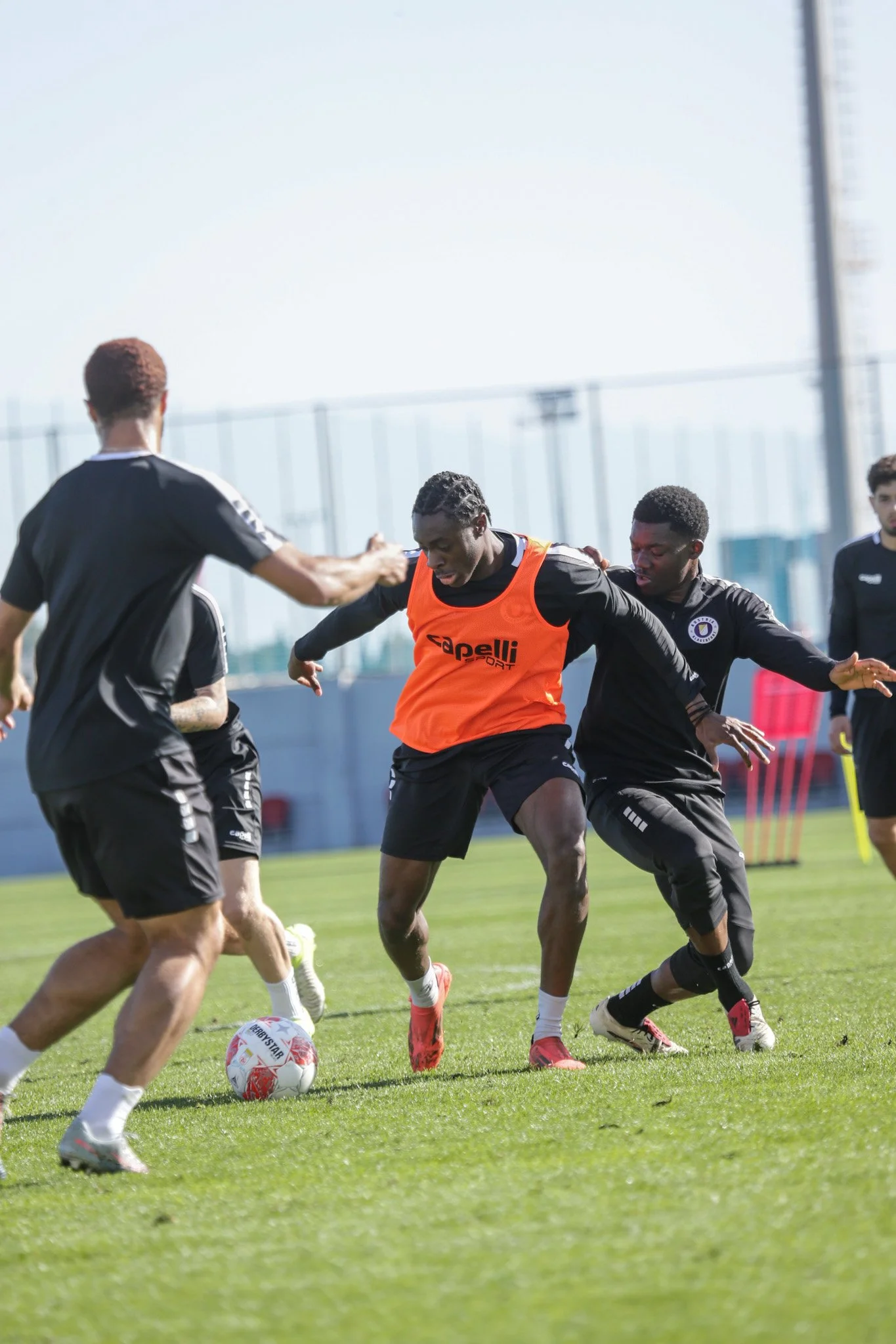 Fußballspieler im Training, mehrere Spieler kämpfen um den Ball auf dem grünen Spielfeld, einige tragen schwarze Trainingskleidung, einer hat eine orange Trainingsweste, es ist tagsüber, im Hintergrund sind Fahnenmaste und Zäune sichtbar.