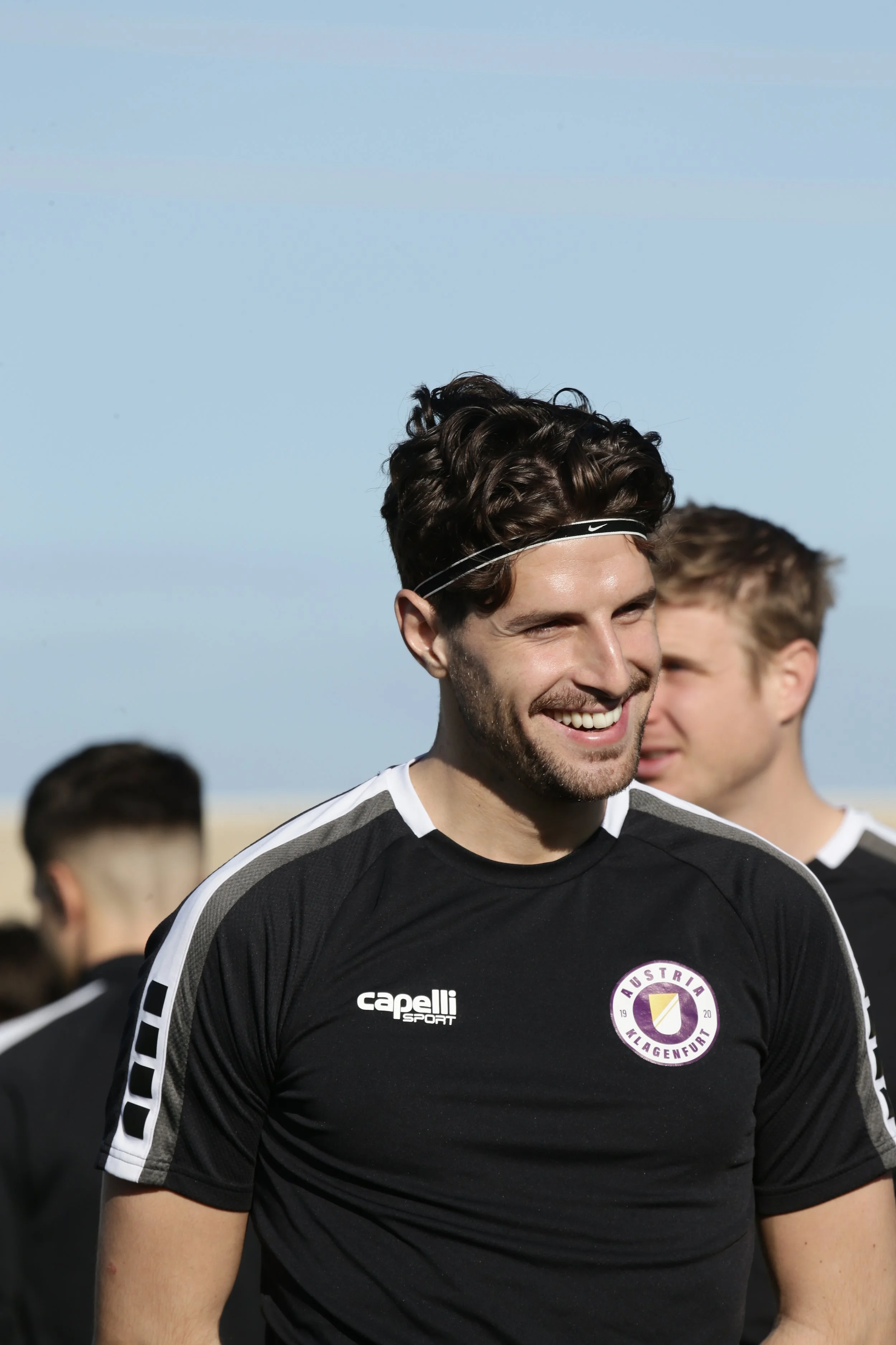 Junger Mann in schwarzem Sports jersey, lächelnd, mit anderen Männern im Hintergrund, auf einem Feld bei Sonnenschein.
