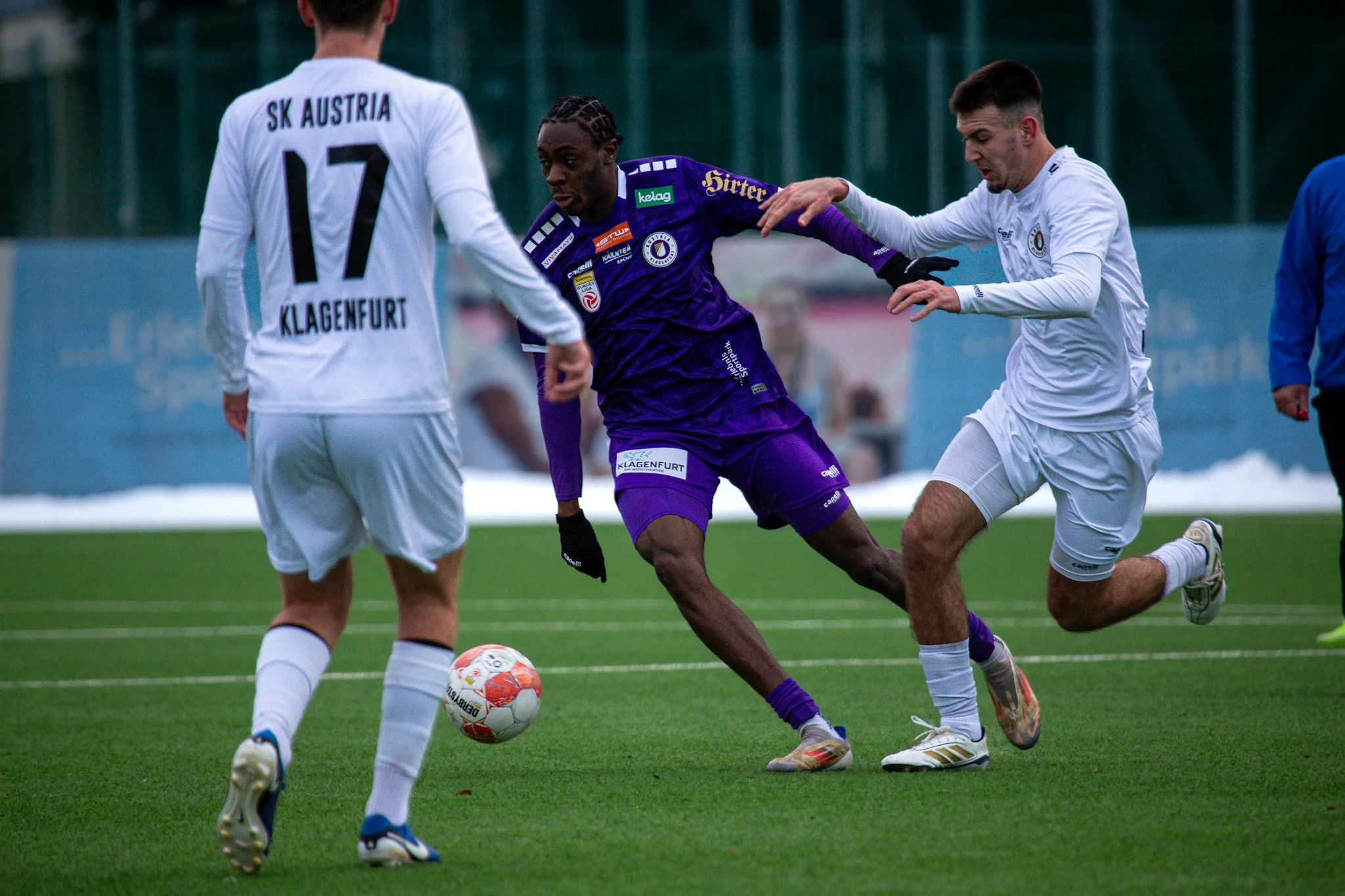 Fußballspieler in Aktion während eines Spiels, mit einem Ball auf einem Fußballfeld, mit Spielern in weißen und lila Trikots.