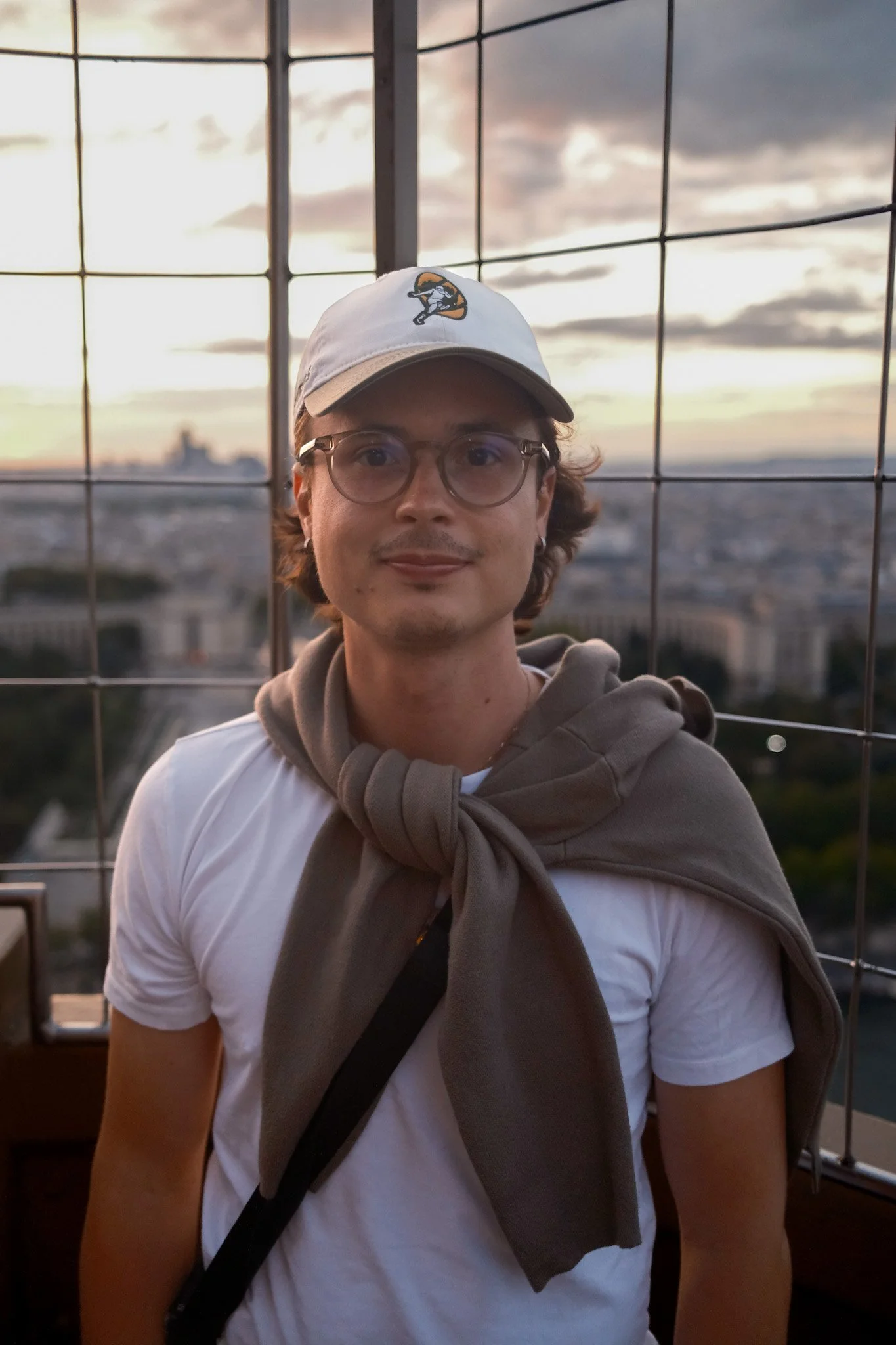 Ein junger Mann mit Brille, weißem T-Shirt, grauer Jacke um den Hals, trägt eine weiße Kappe mit einem Logo, steht vor einer Gitterabsperrung bei Sonnenuntergang, mit Blick auf die Stadt Paris im Hintergrund.