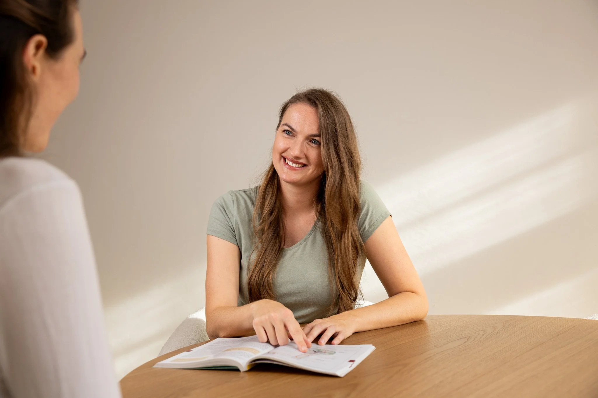 Zwei Frauen sitzen an einem runden Holztisch. Die Frau auf der rechten Seite, die sichtbar ist, lächelt und zeigt auf eine offene Zeitschrift, die auf dem Tisch liegt. Die andere Frau, nur teilweise sichtbar, sitzt gegenüber.