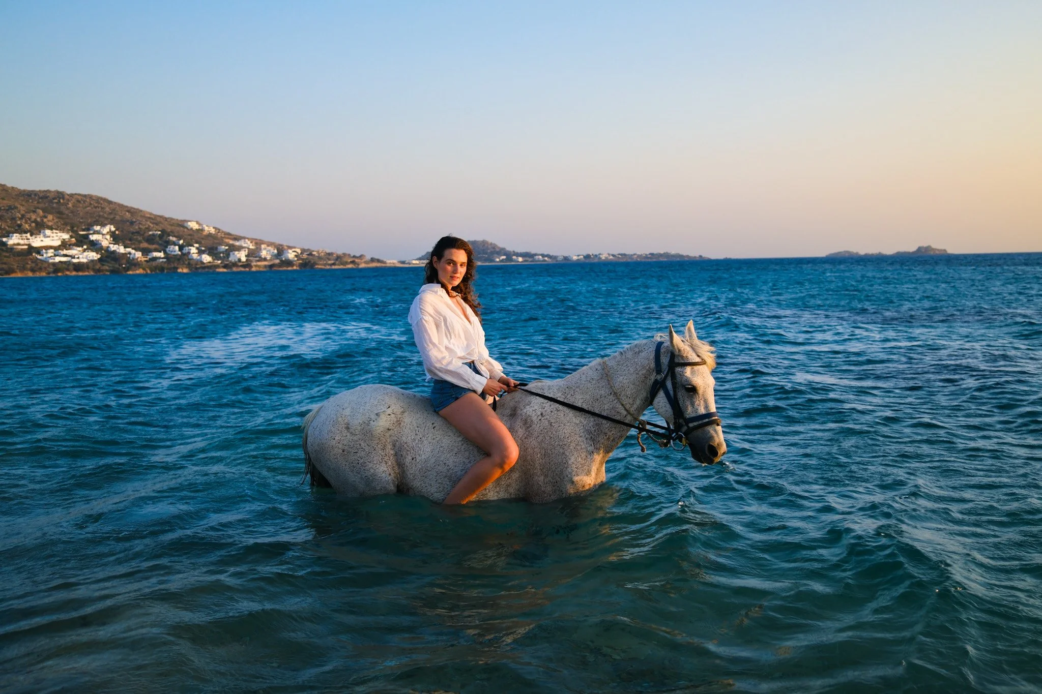 Frau reitet auf einem weißen Pferd im Wasser nahe der Küste bei Sonnenuntergang.
