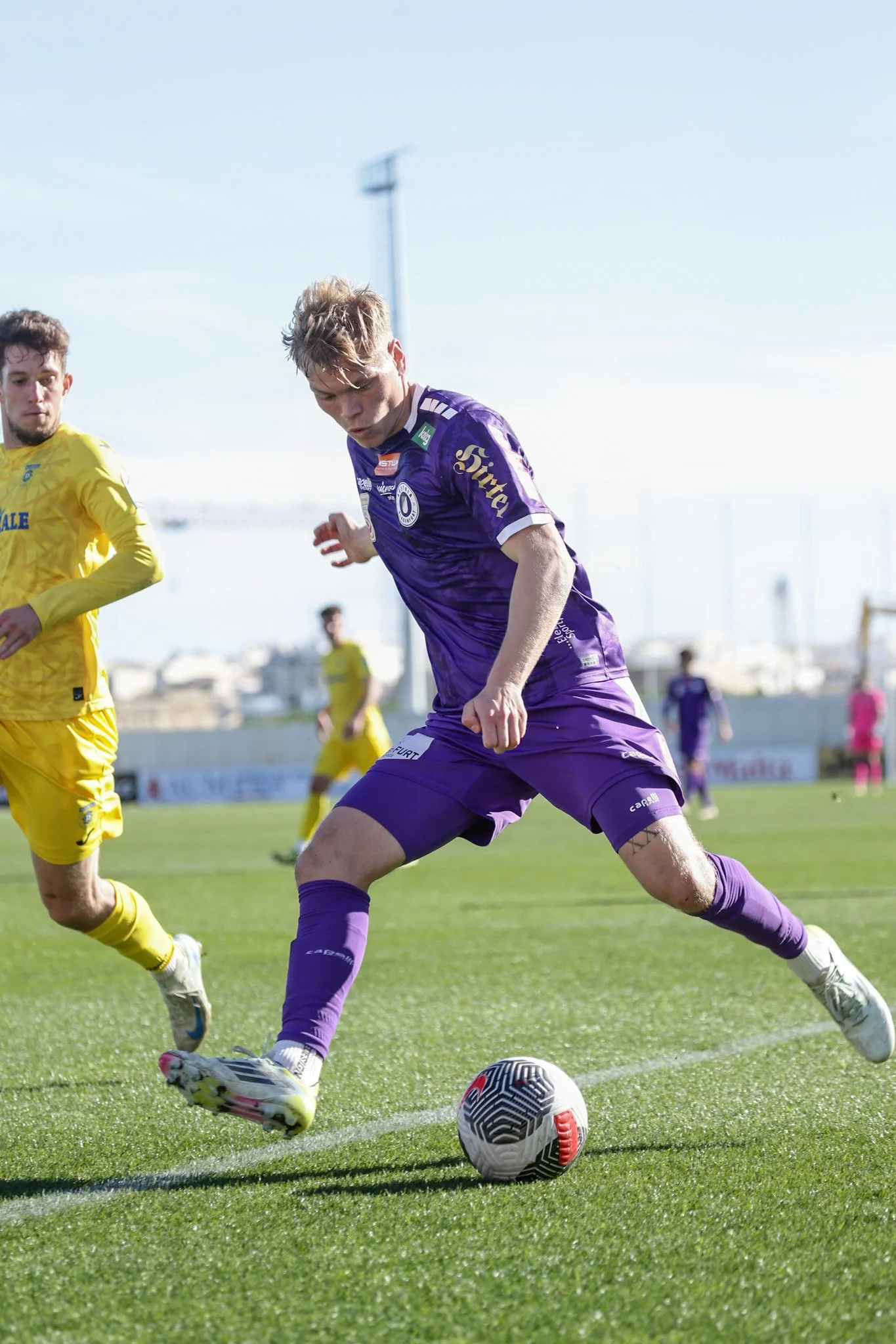 Fußballspieler in lila Trikot und Shorts, der den Ball auf dem Spielfeld kontrolliert, während ein Gegenspieler in gelbem Trikot und Shorts daneben läuft.