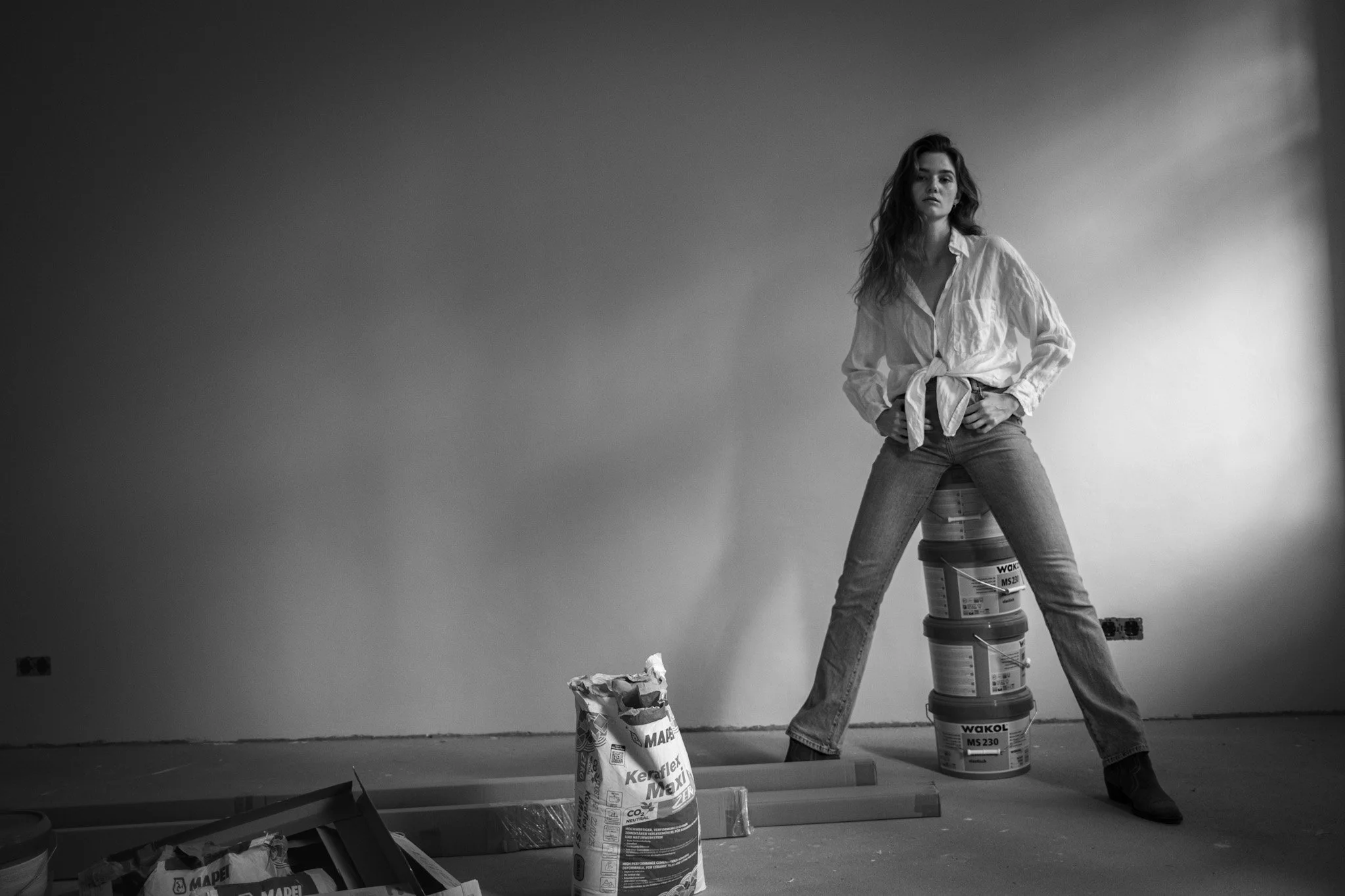 Eine junge Frau steht in einem Raum mit einer leeren Wand. Sie trägt eine offene Bluse, die vorne geknüpft ist, und Jeans. Sie steht mit den Beinen breit auseinander, auf einem Stapel von Farbdosen, und blickt ernst in die Kamera.