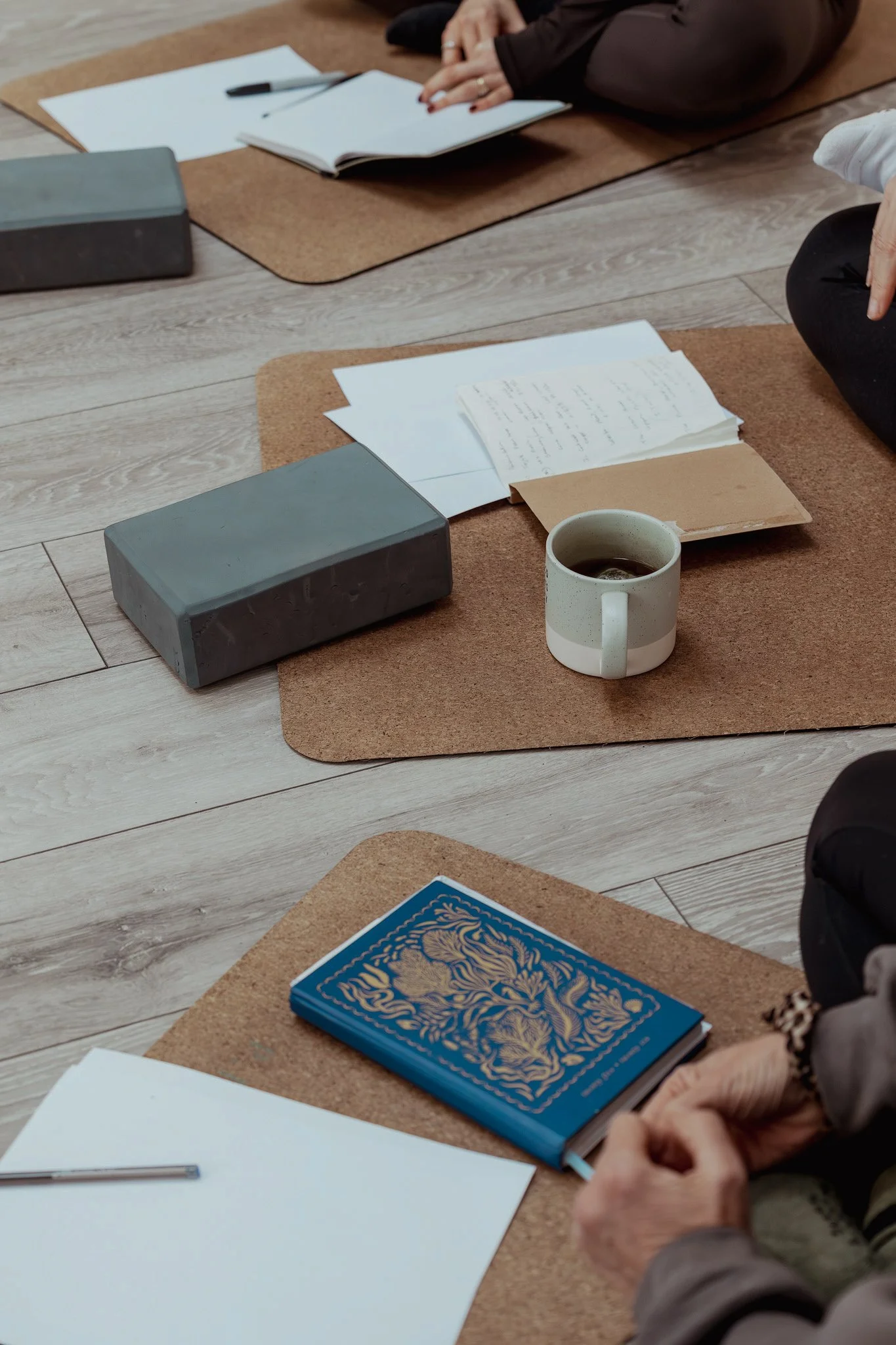 A person seated cross-legged on the floor with their hands clasped, surrounded by books, papers, a coffee mug, and blocks on cork mats on a wooden floor.