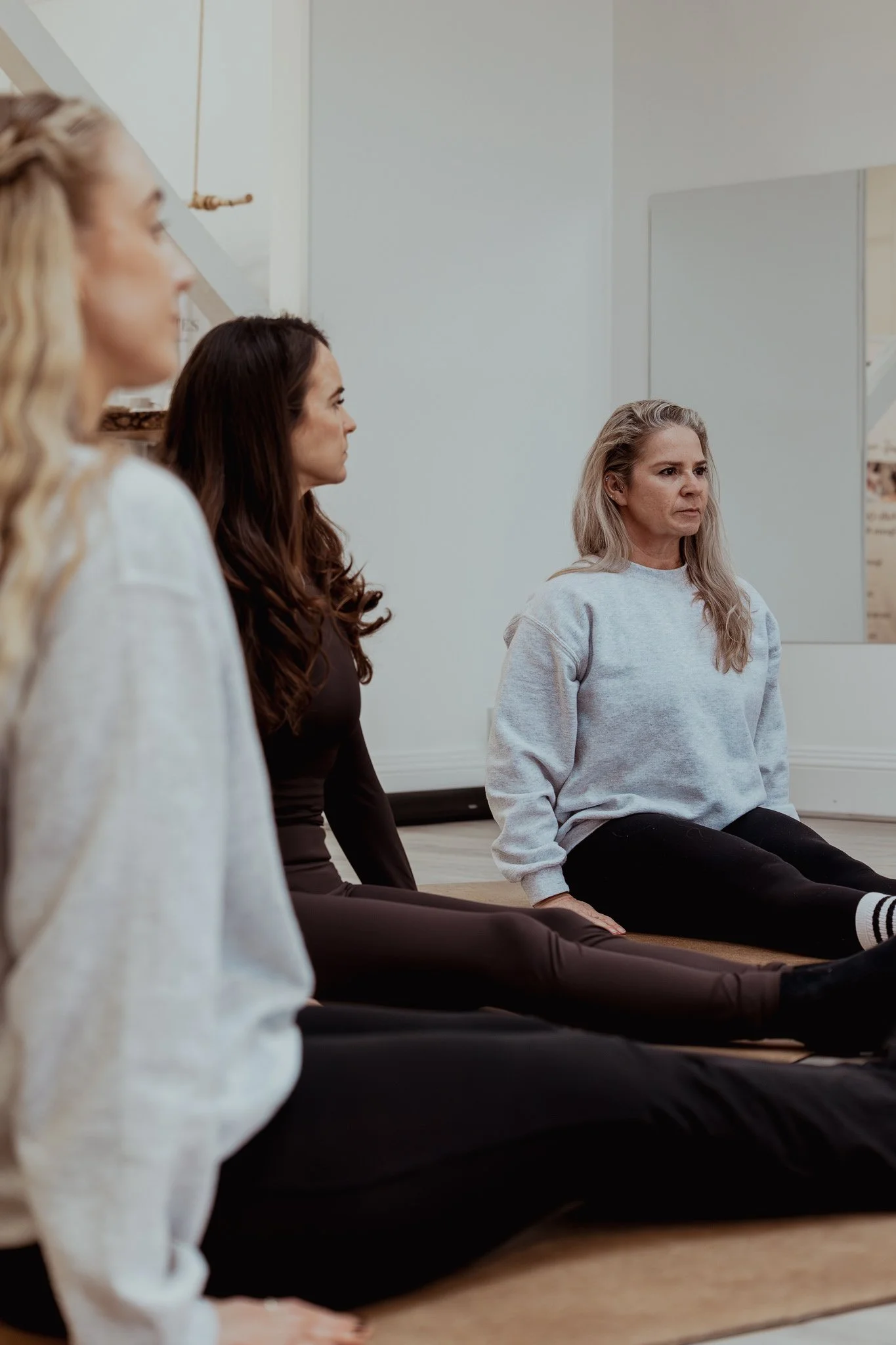 Three women sitting on the floor in a yoga studio, practicing yoga or meditation. They are facing a mirror and sitting in a cross-legged position.