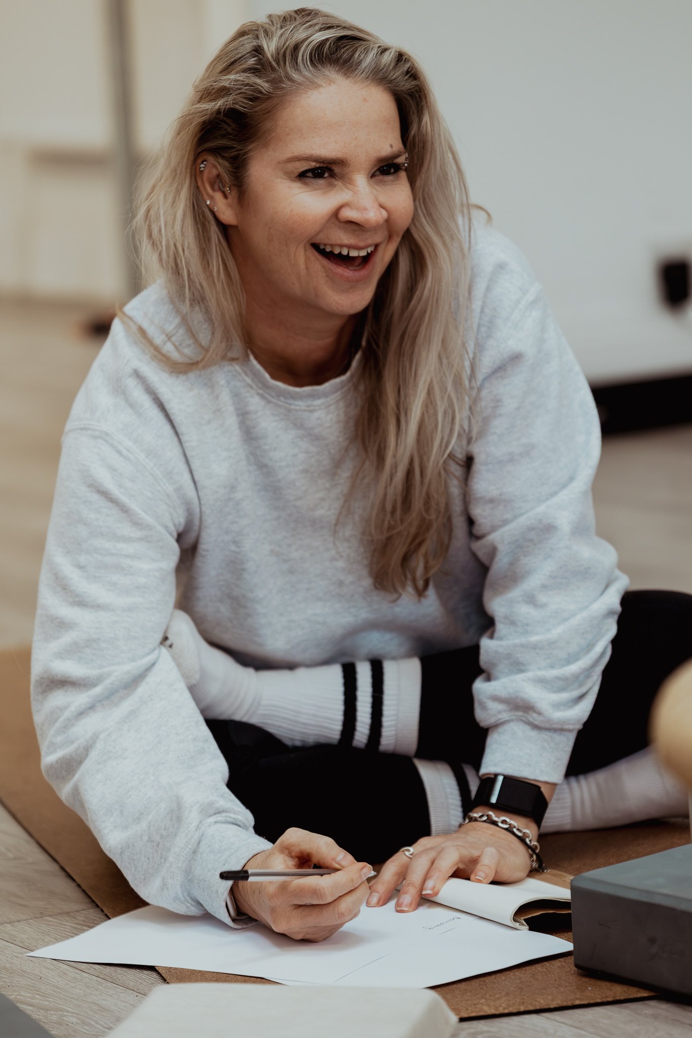 A young woman with long, blonde hair, wearing a gray sweatshirt and black-and-white striped socks, is sitting on the floor, smiling, and writing in a notebook.