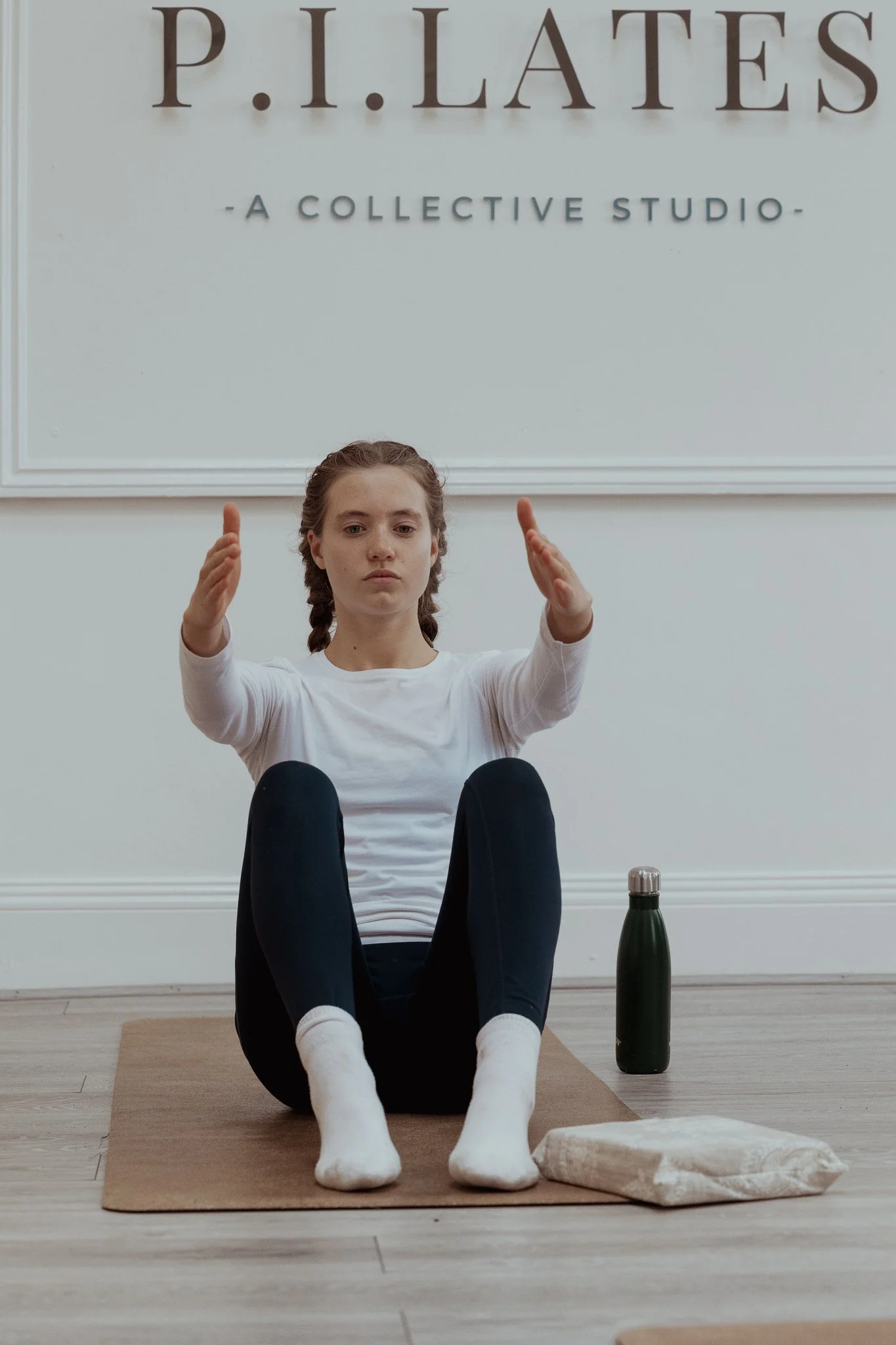 A young girl practicing a seated yoga pose on a yoga mat inside a studio. She is wearing a white long-sleeve shirt, black leggings, and white socks. A water bottle and a folded towel are placed beside her. A sign in the background reads "P.I. LATES - A COLLECTIVE STUDIO."